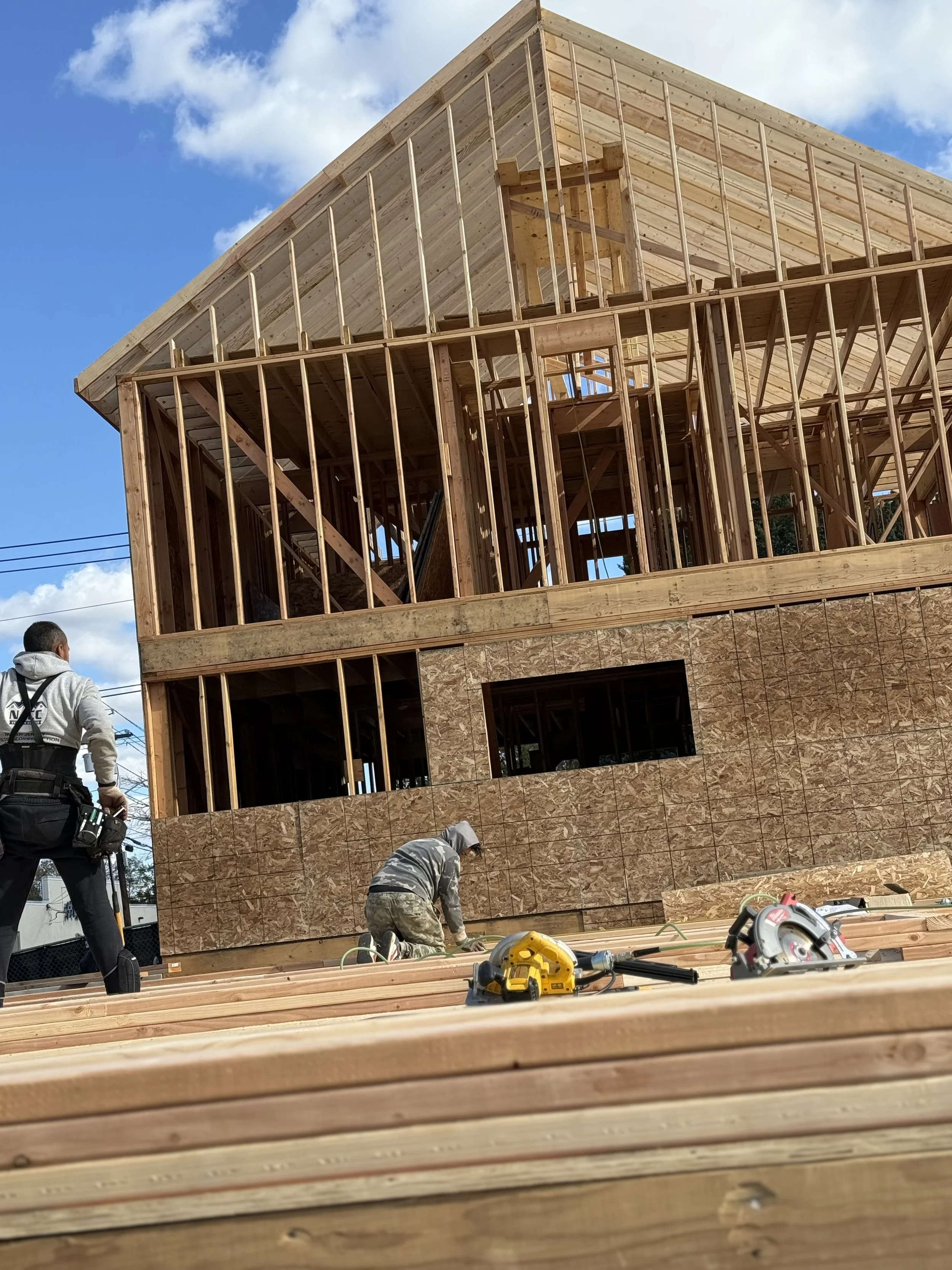 Building under construction with wooden framing and plywood sheathing, construction workers working on the site, construction tools on the foreground, blue sky with some clouds