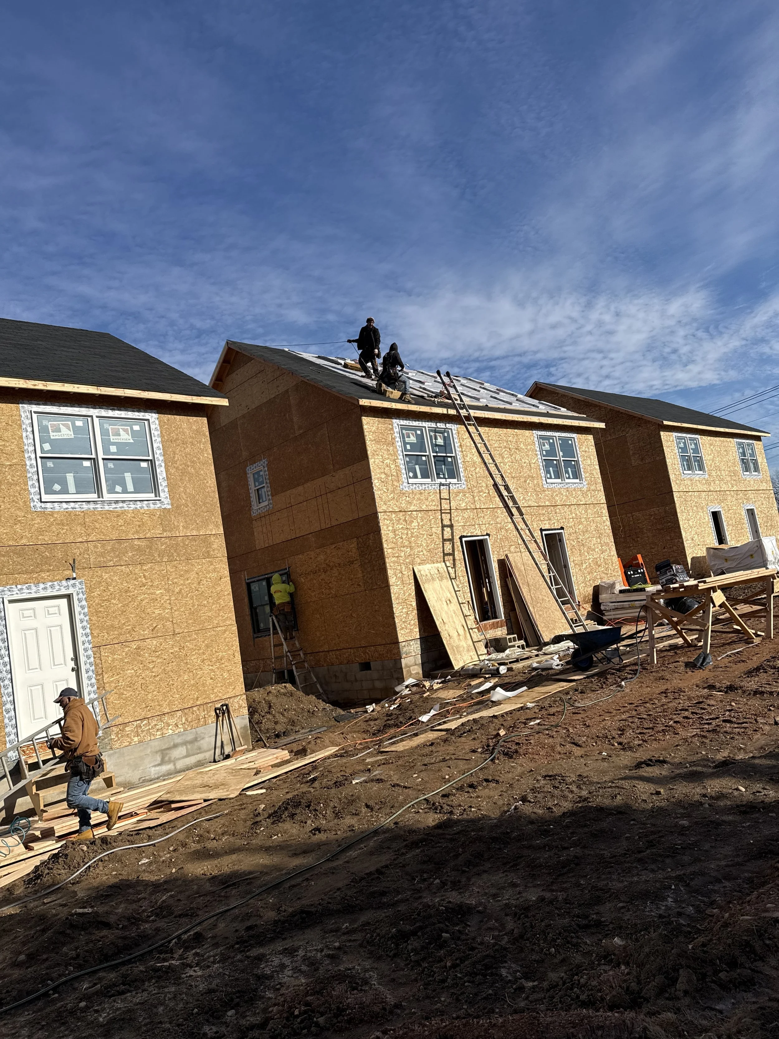 Construction workers building a house, working on the roof and walls, surrounded by tools and materials, in a clear outdoor setting. Framing contractors framing 3 homes on a sunny day.