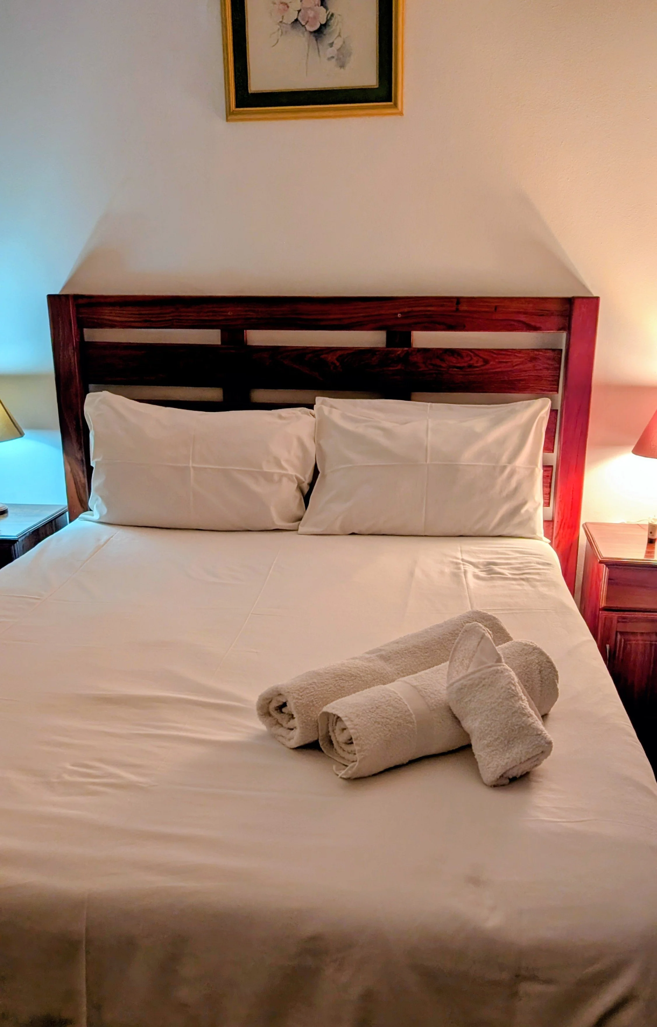 A neatly made hotel bed with white linens, three rolled white towels on top, and wooden headboard. Two pillows are arranged against the headboard. There are two bedside tables with lamps on each side, and a framed floral picture hangs above the bed.