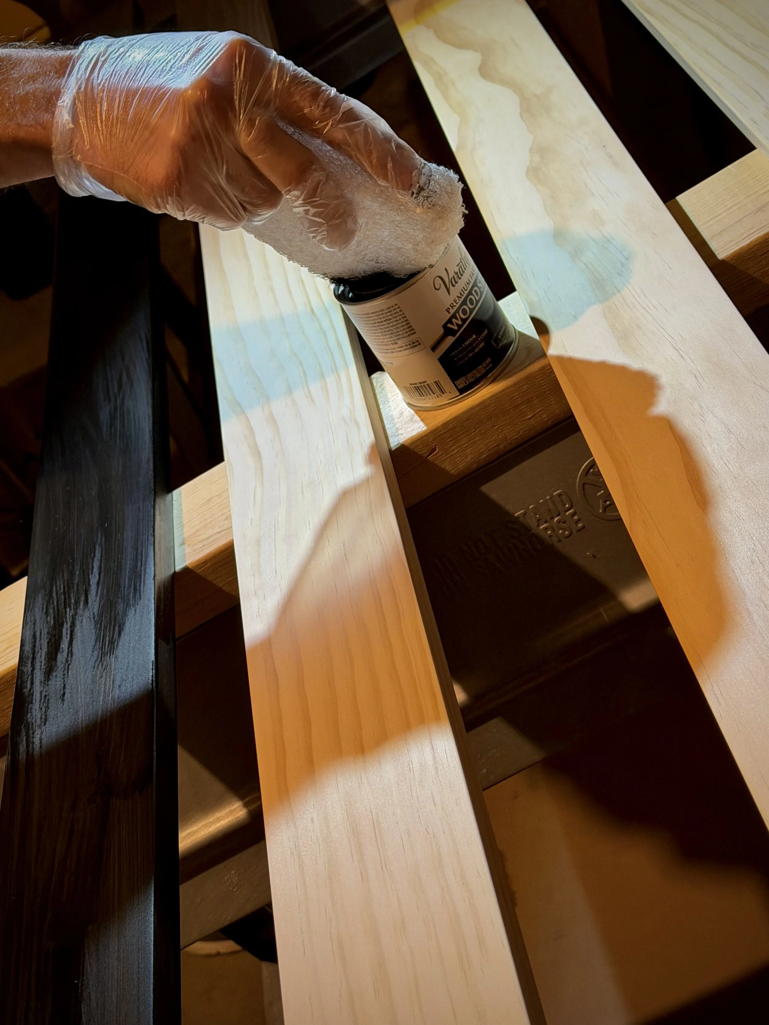 Person wearing a clear glove applying stain or finish to a wooden bench with a rag, with the bench resting on a work surface.