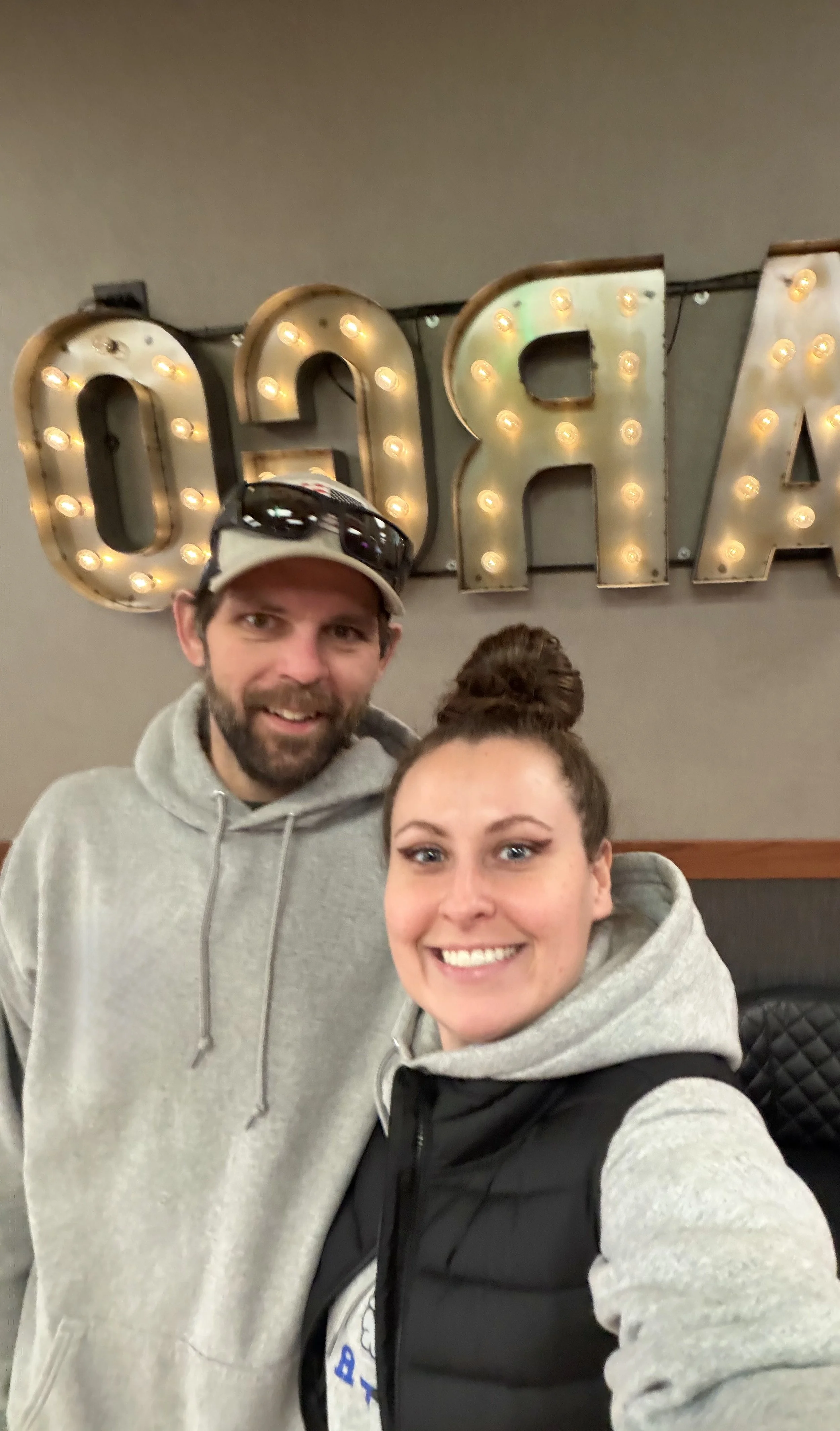 A man and a woman taking a selfie in front of a large illuminated sign that spells out 'OAR'. The man is wearing sunglasses on his cap and a gray hoodie, while the woman has her hair in a bun and is wearing a gray hoodie with a black vest.