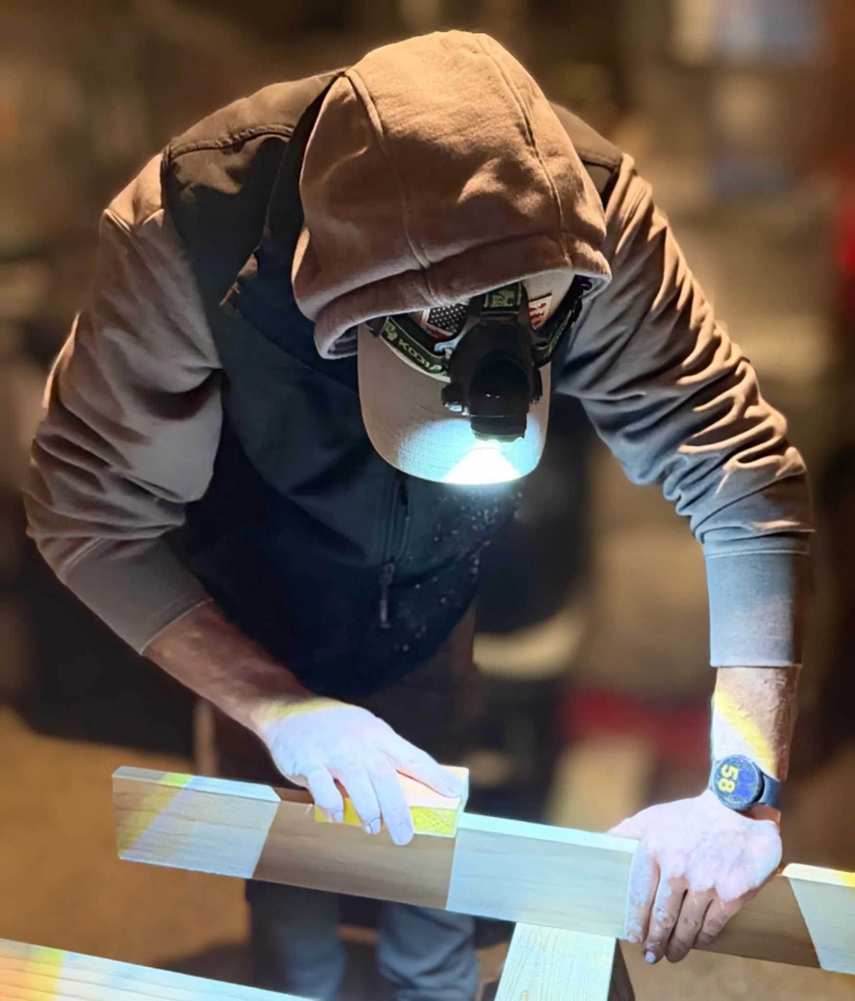 A person wearing a hoodie, cap, gloves, and a headlamp is working on a piece of wood in a workshop.