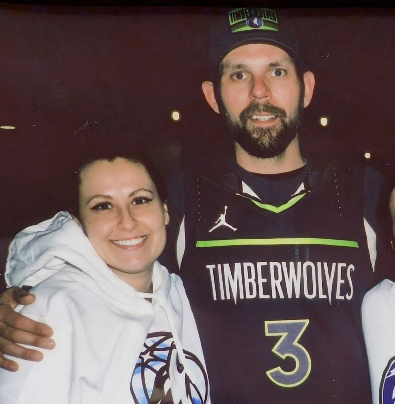 A woman smiling and wearing a white hoodie with a basketball logo, standing next to a man in a Timberwolves basketball jersey with the number 3. They are indoors with dim lighting.