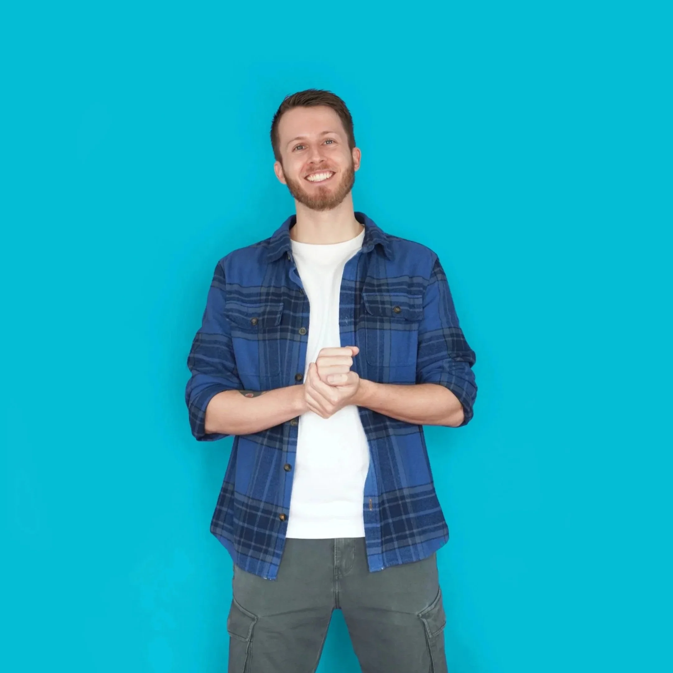 A young man with short brown hair and a beard smiling against a bright blue background, wearing a blue plaid shirt over a white T-shirt and gray jeans.