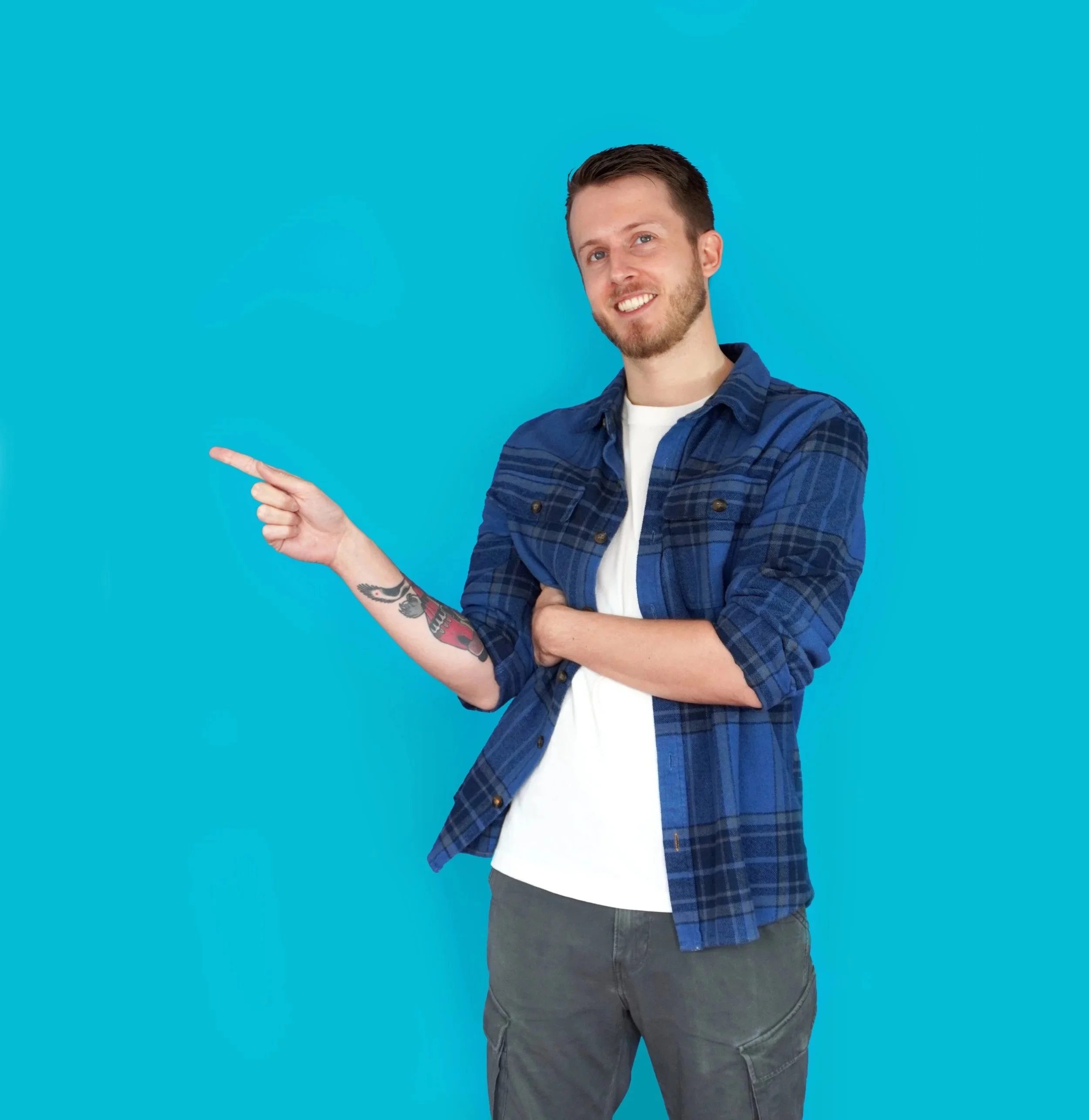 A young man with short dark hair and a beard wearing a blue plaid shirt and gray pants, smiling and pointing to his left against a bright blue background.