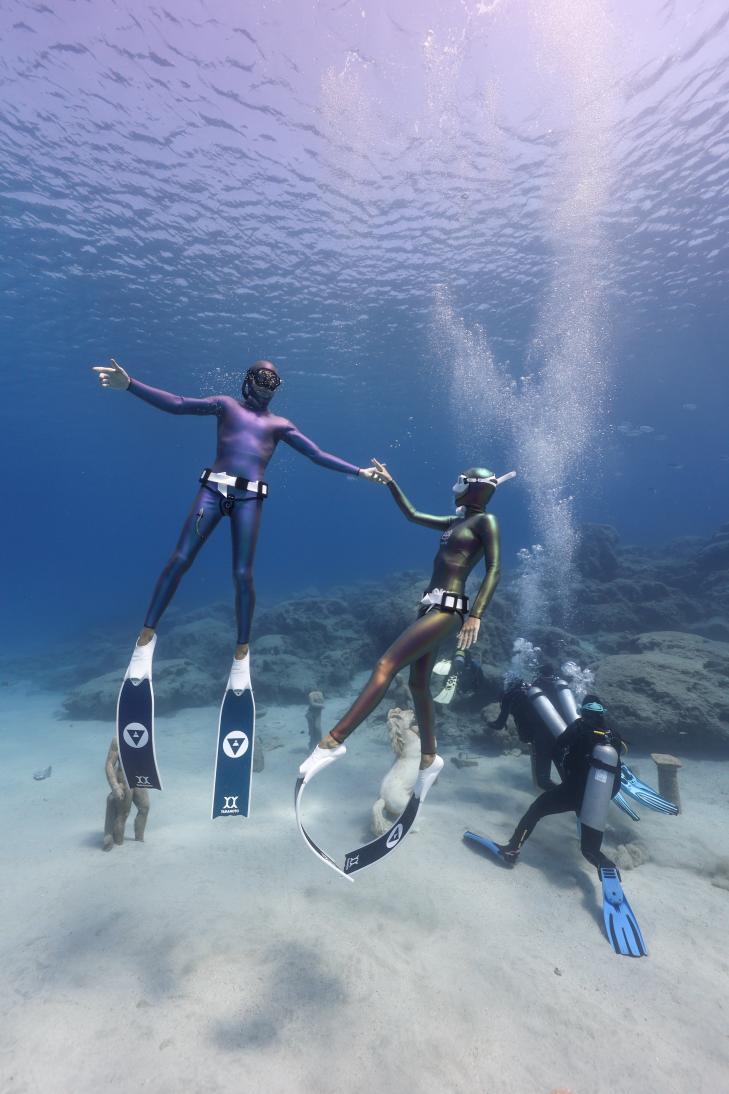 Two freedivers holding hands underwater near scuba divers exploring the seabed in Protaras, Cyprus, illustrating the contrast between freediving and scuba diving.