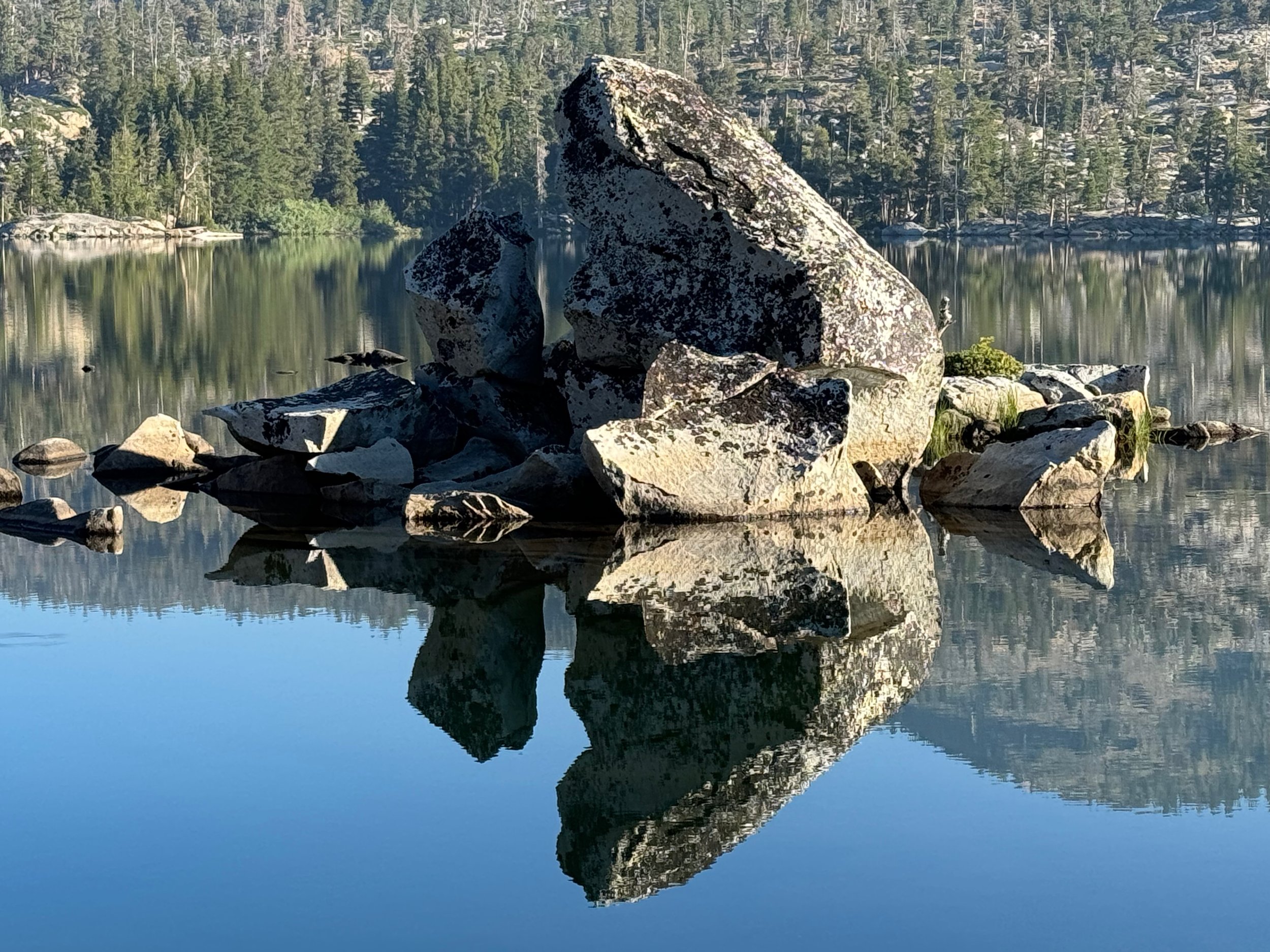 Large rocks and boulders in a calm lake with forested shoreline and mountains reflected in the water.