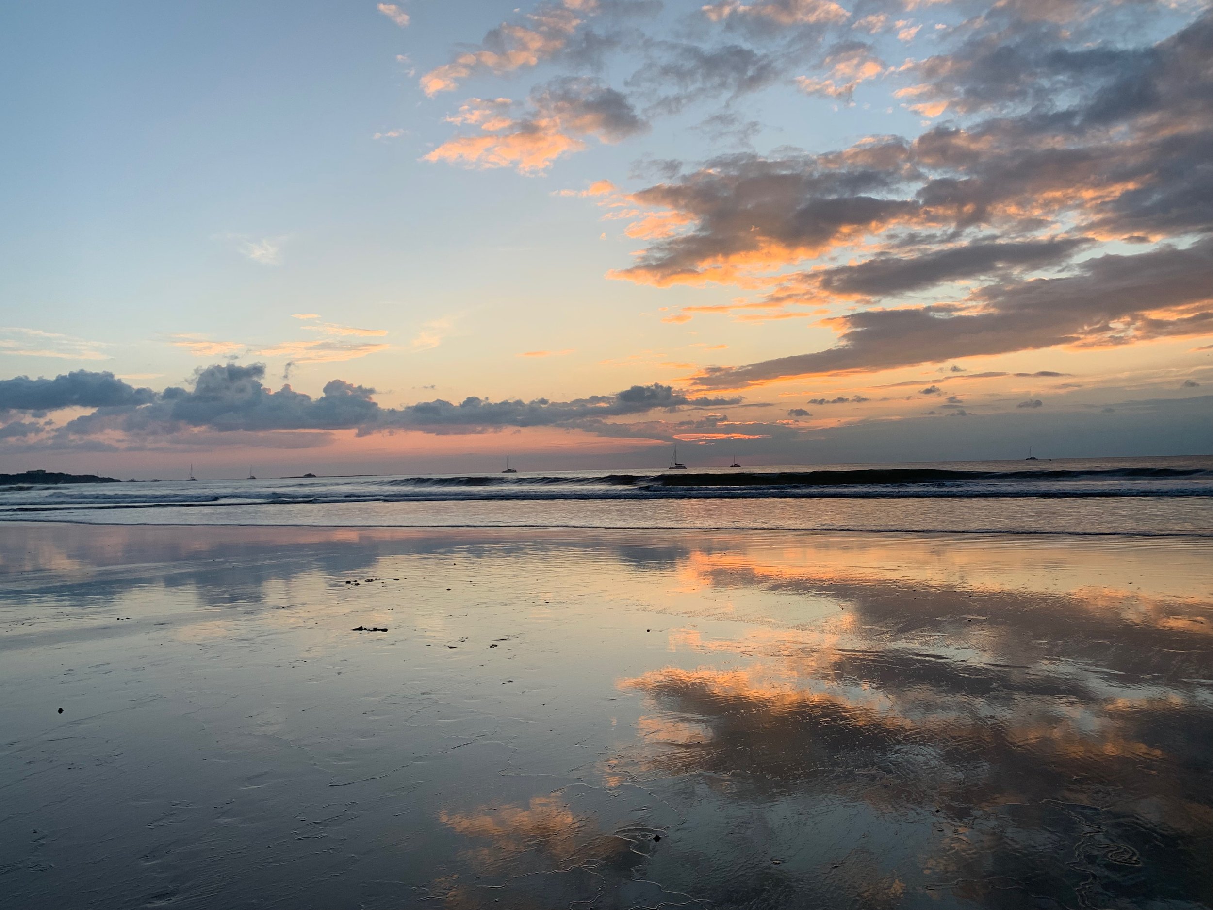 Sunset over a calm beach with clouds reflected on the wet sand and the ocean in the background.