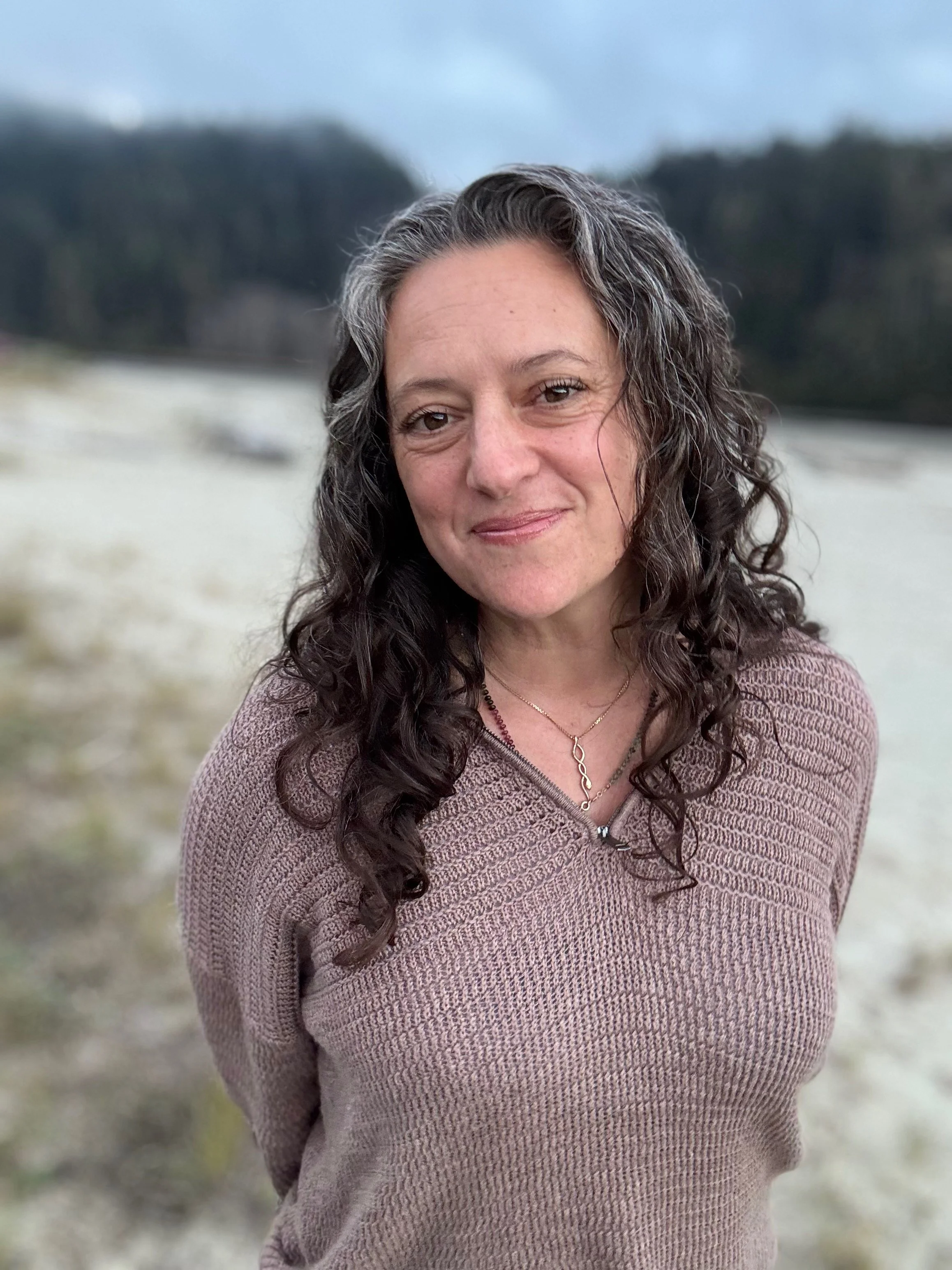 A woman with long, curly dark hair and light skin standing outdoors with a blurred natural landscape in the background.