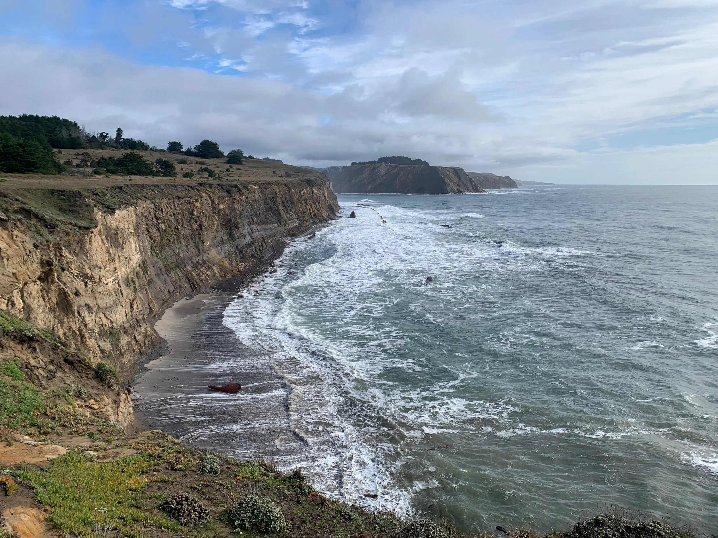 Coastal cliffs with grassy top area, rocky shoreline, and waves crashing against the coast under a partly cloudy sky.