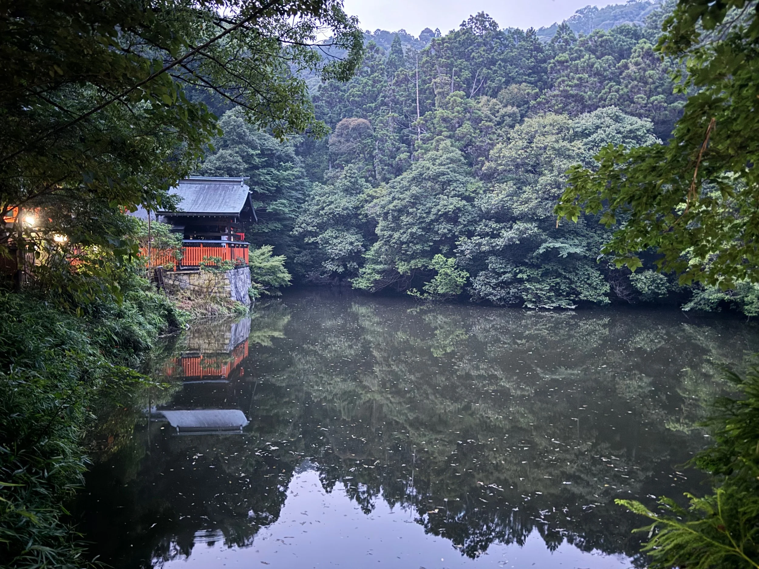 A serene lake surrounded by dense green trees with a traditional Japanese wooden building on the left side, reflected in the water.