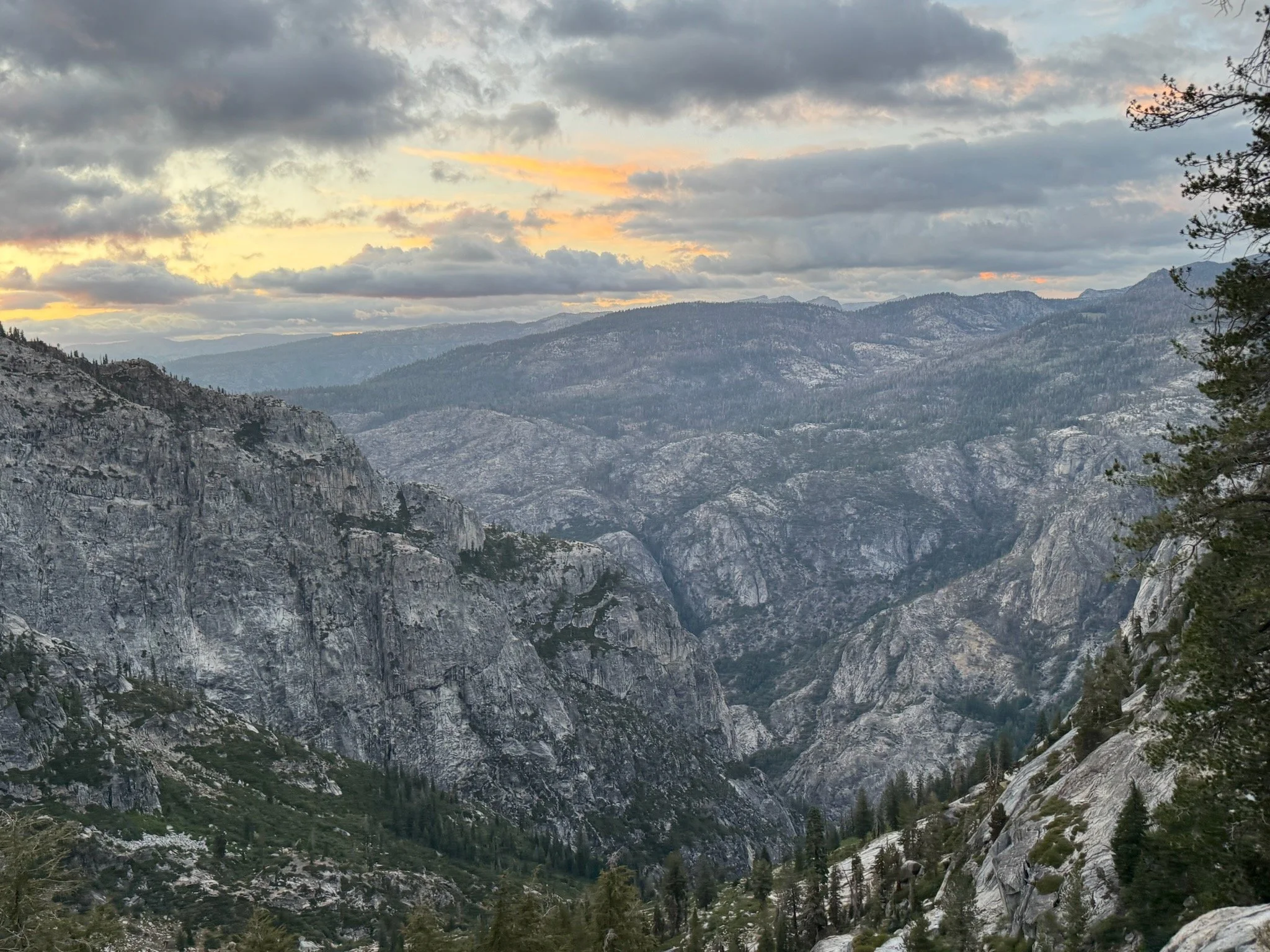 A rugged mountain landscape at sunset with a cloudy sky, featuring steep rocky cliffs, wooded slopes, and distant mountain ridges. Yosemite National Park