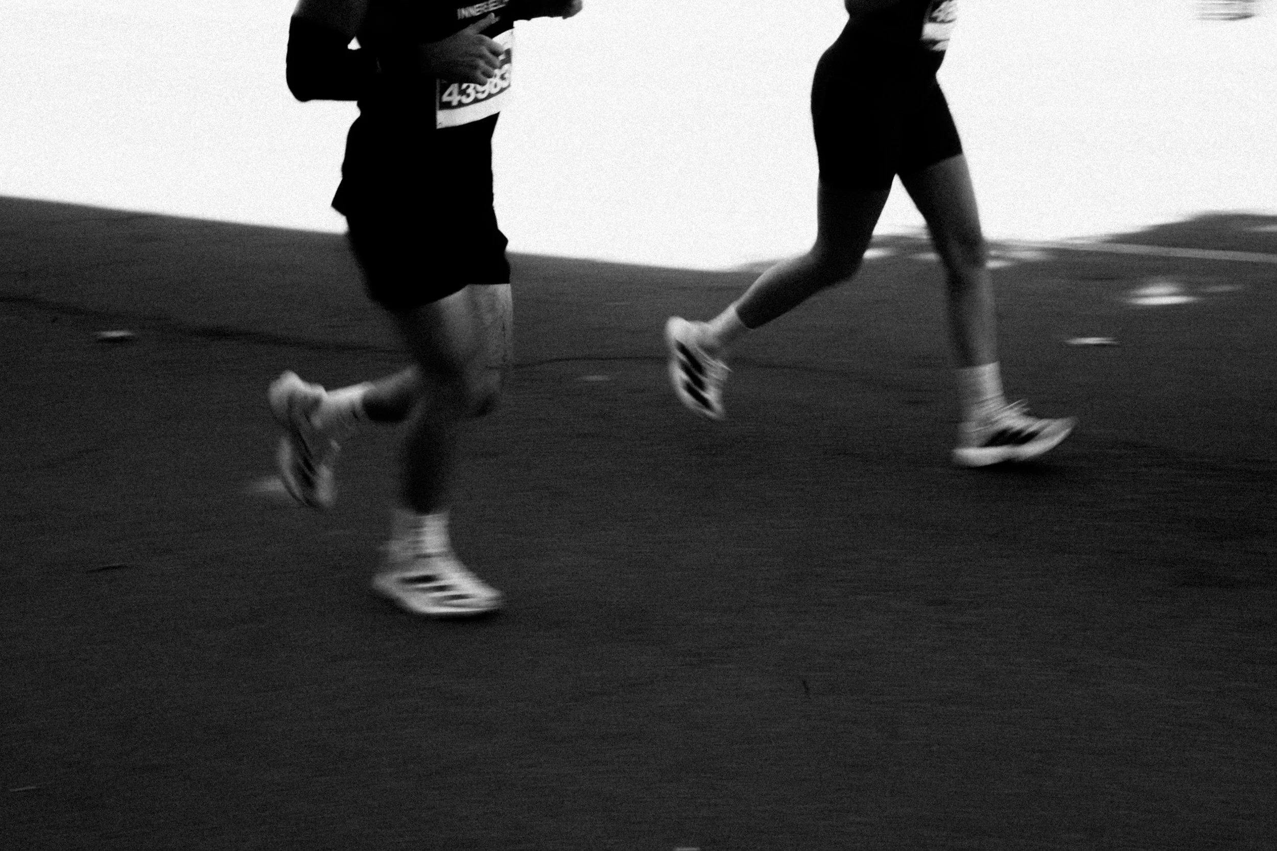 Black and white photo of two runners jogging indoors, showing their legs and feet, with one running in a sports shoe, and a wall in the background.