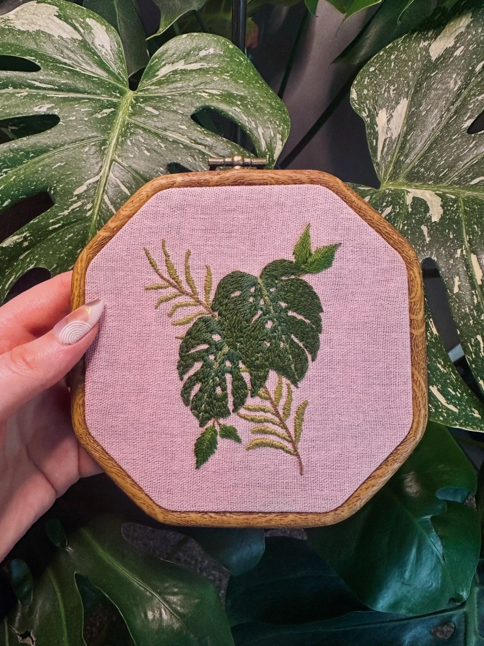 Embroidery of green leaves on pink fabric held in a wooden hoop, with Monstera plant leaves in the background.