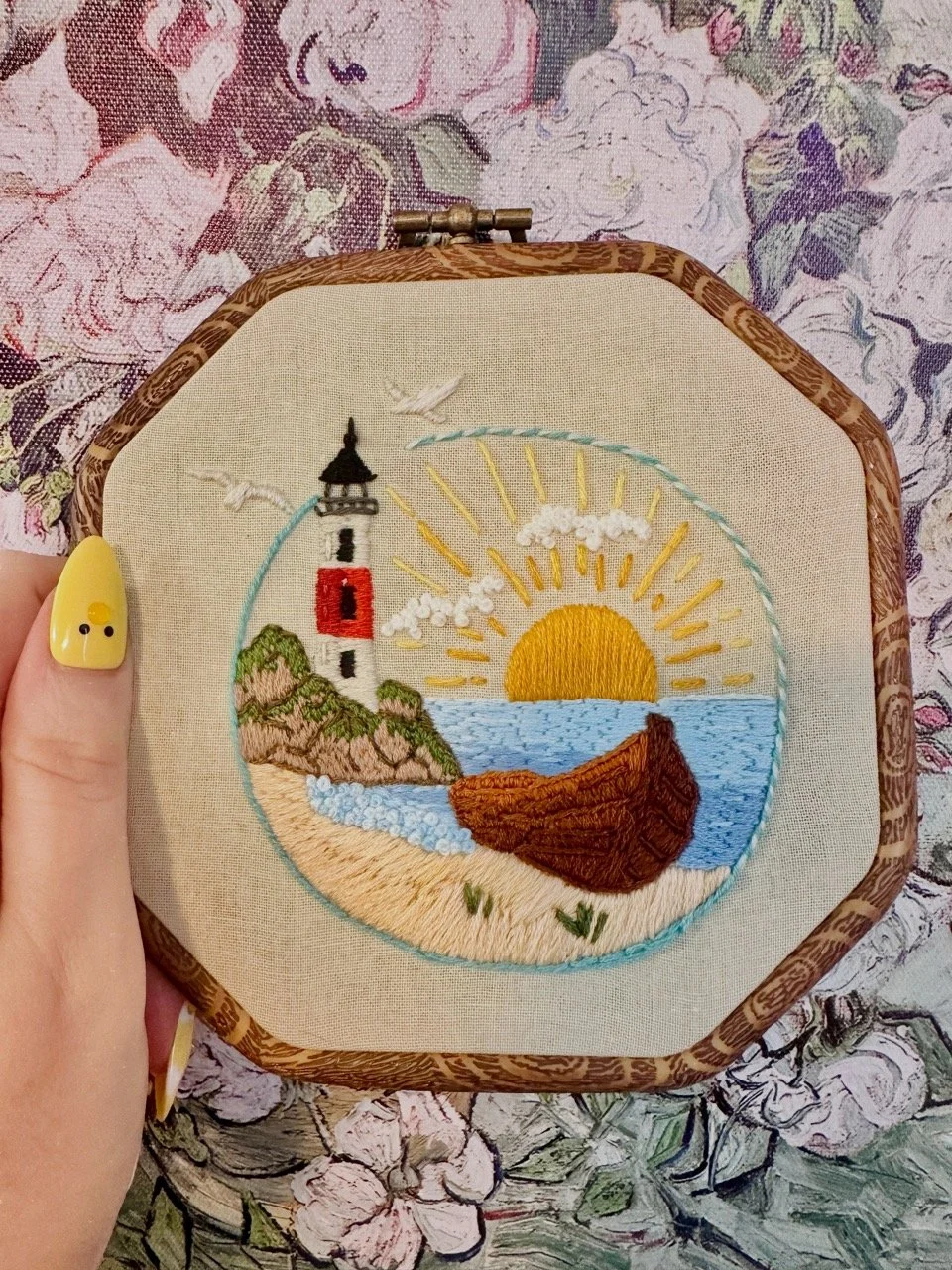 Embroidery of a beach scene with a lighthouse, sun setting over the ocean, sandy shore, and rocks, in a wooden hoop