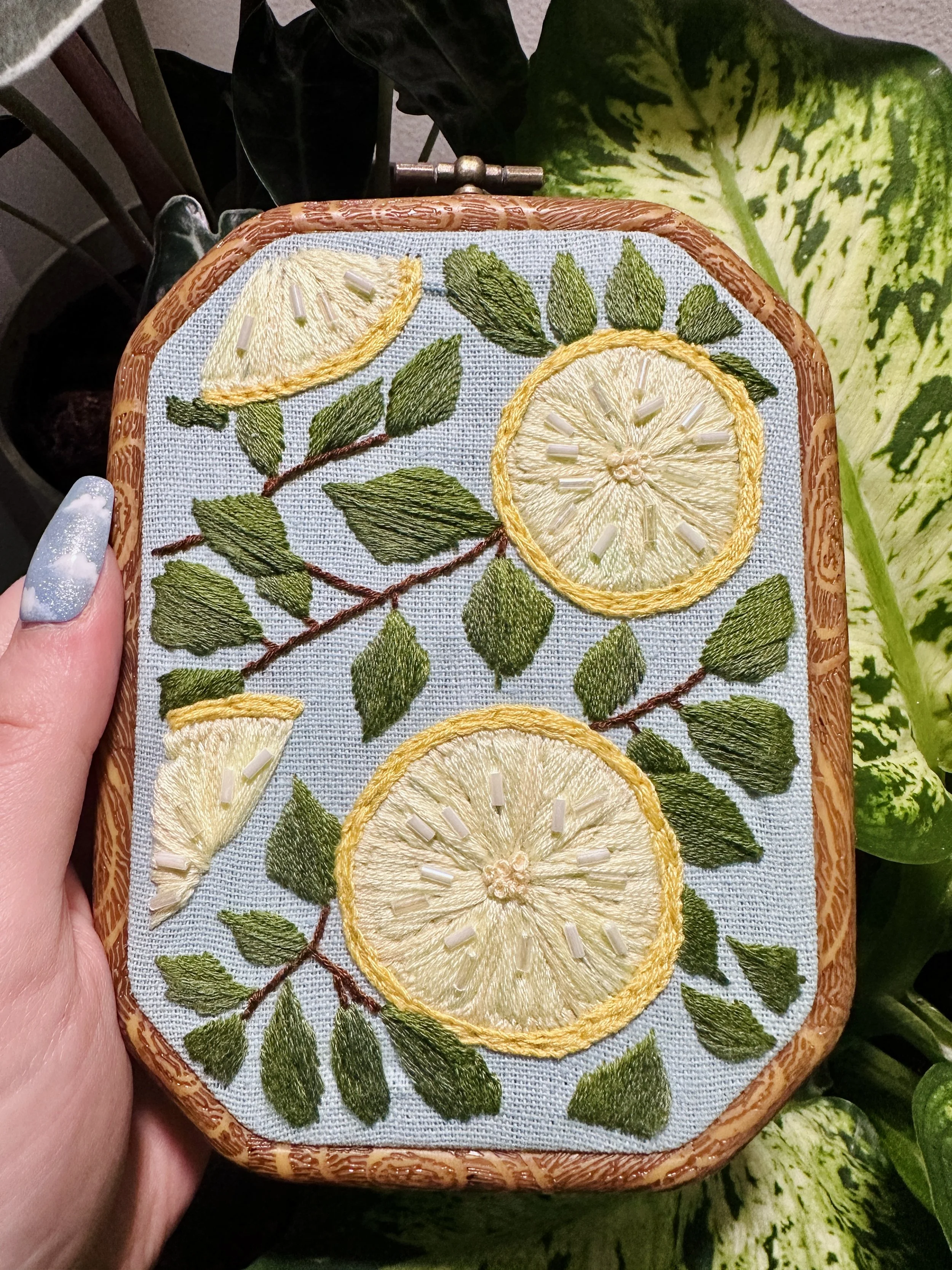 Embroidery of lemon slices and leaves on fabric held in a hand with painted nails.