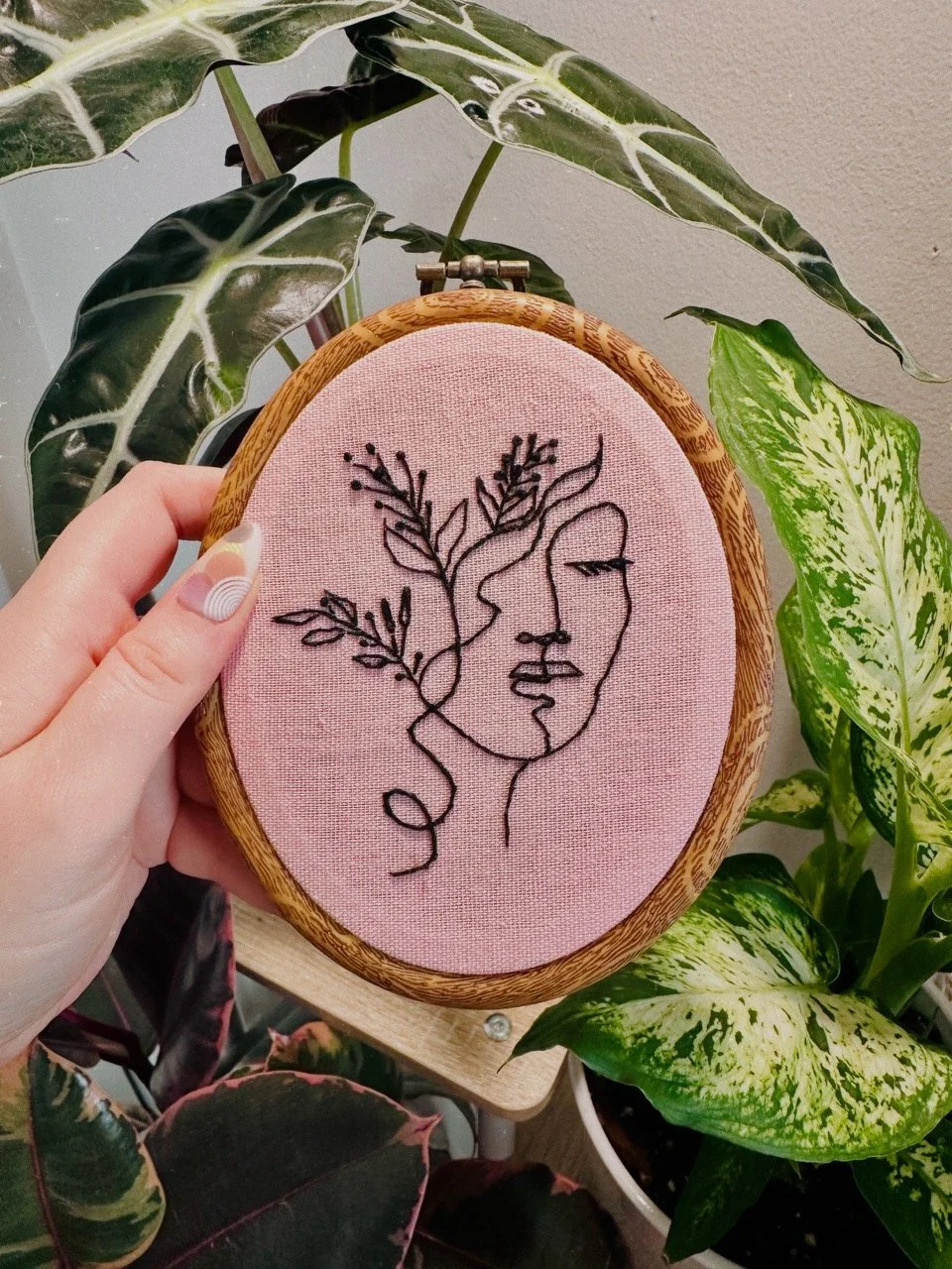 A hand holding an embroidery hoop with pink fabric, featuring a black line art of a woman's face intertwined with leafy branches, surrounded by green houseplants.