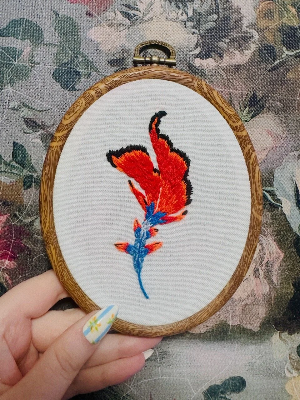 Embroidery of a red fox with a blue tail on white fabric, held in a wooden hoop, against a floral fabric background.