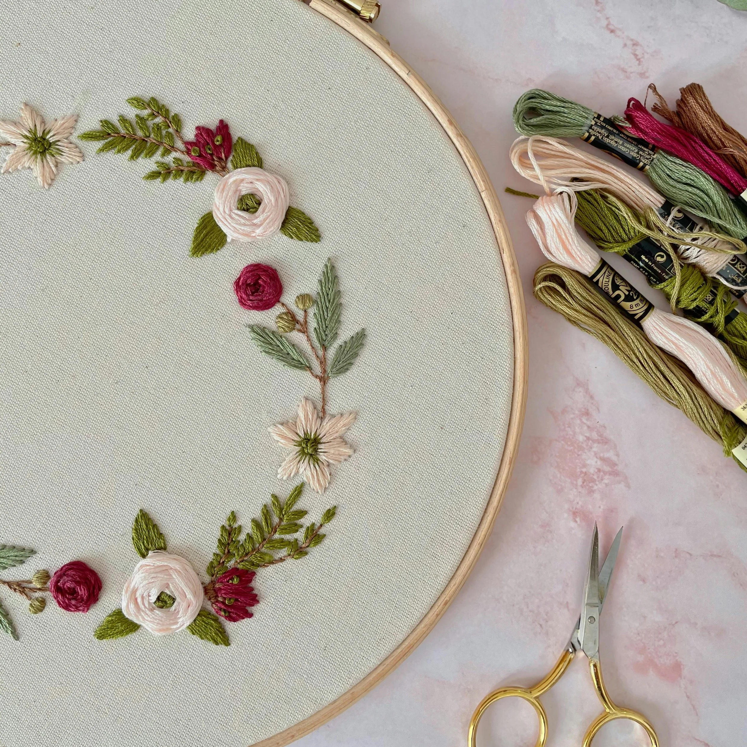 Embroidery hoop with floral embroidery, embroidery threads, scissors, and a pink marbled background.