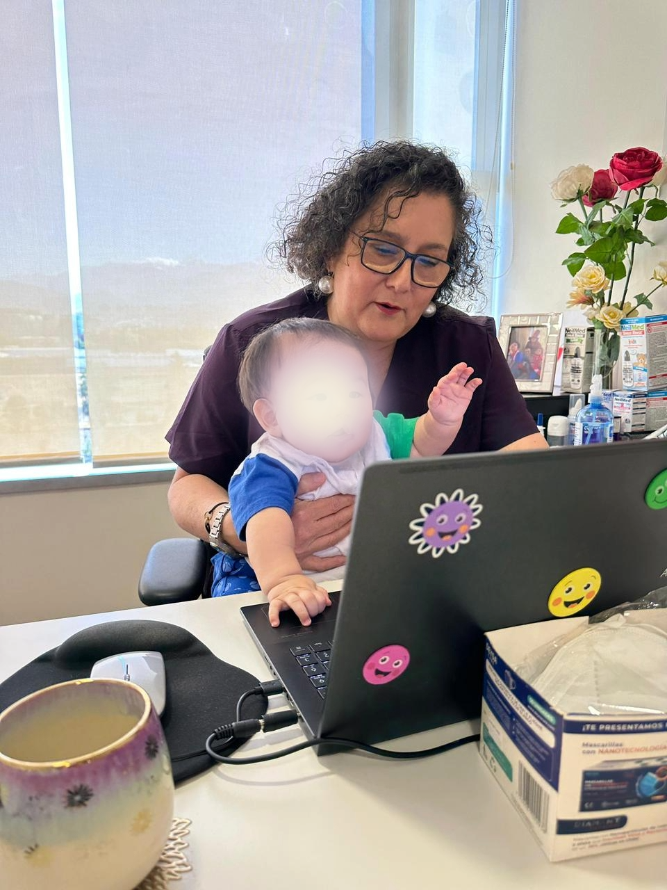 Una mujer con gafas y peinado rizado, con aretes grandes, está sentada en una oficina con un niño en su regazo, ambos frente a una laptop decorada con pegatinas de caras felices y animales. La mujer parece estar explicando algo al niño, que mira la pantalla. Detrás hay una ventana, una mesa con medicamentos, un marco con una foto y un florero con rosas.