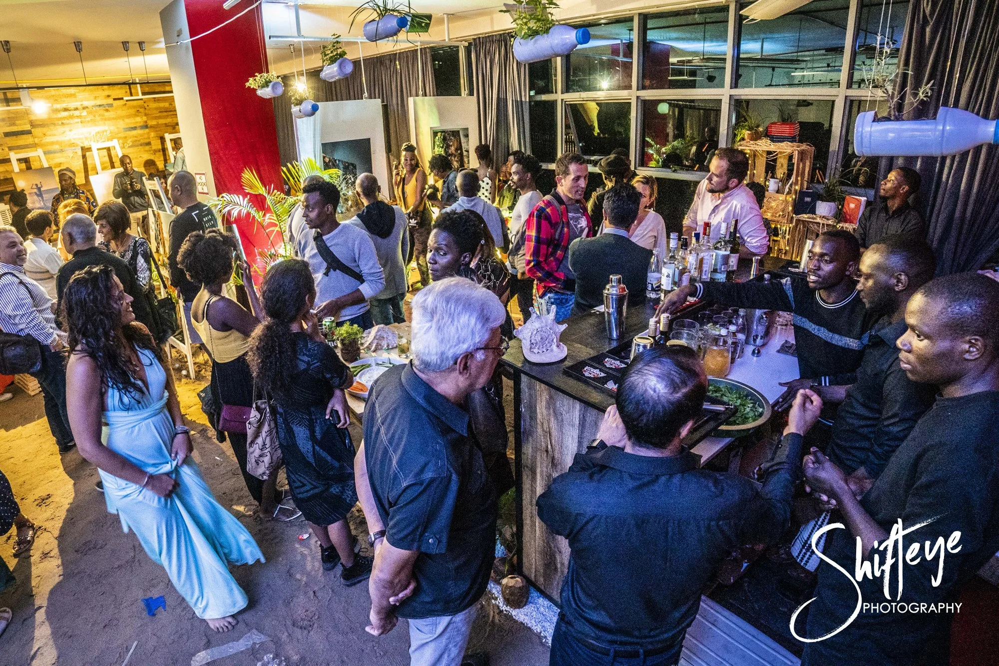 A crowded indoor art event or gallery reception with diverse attendees, some viewing artwork, others socializing, with a bar area serving drinks, and art pieces displayed on easels and walls.