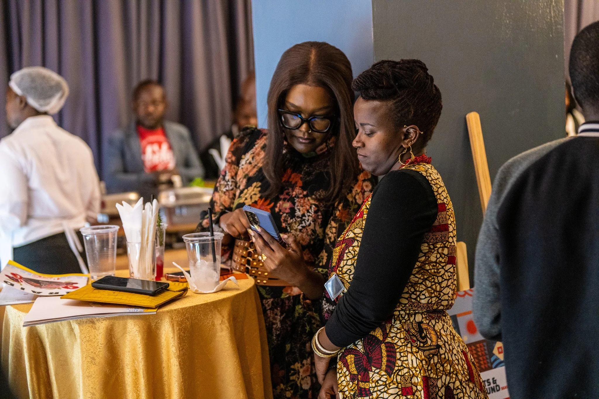 Two women standing at a table, looking at a smartphone together at an indoor event, with other people and event materials in the background.
