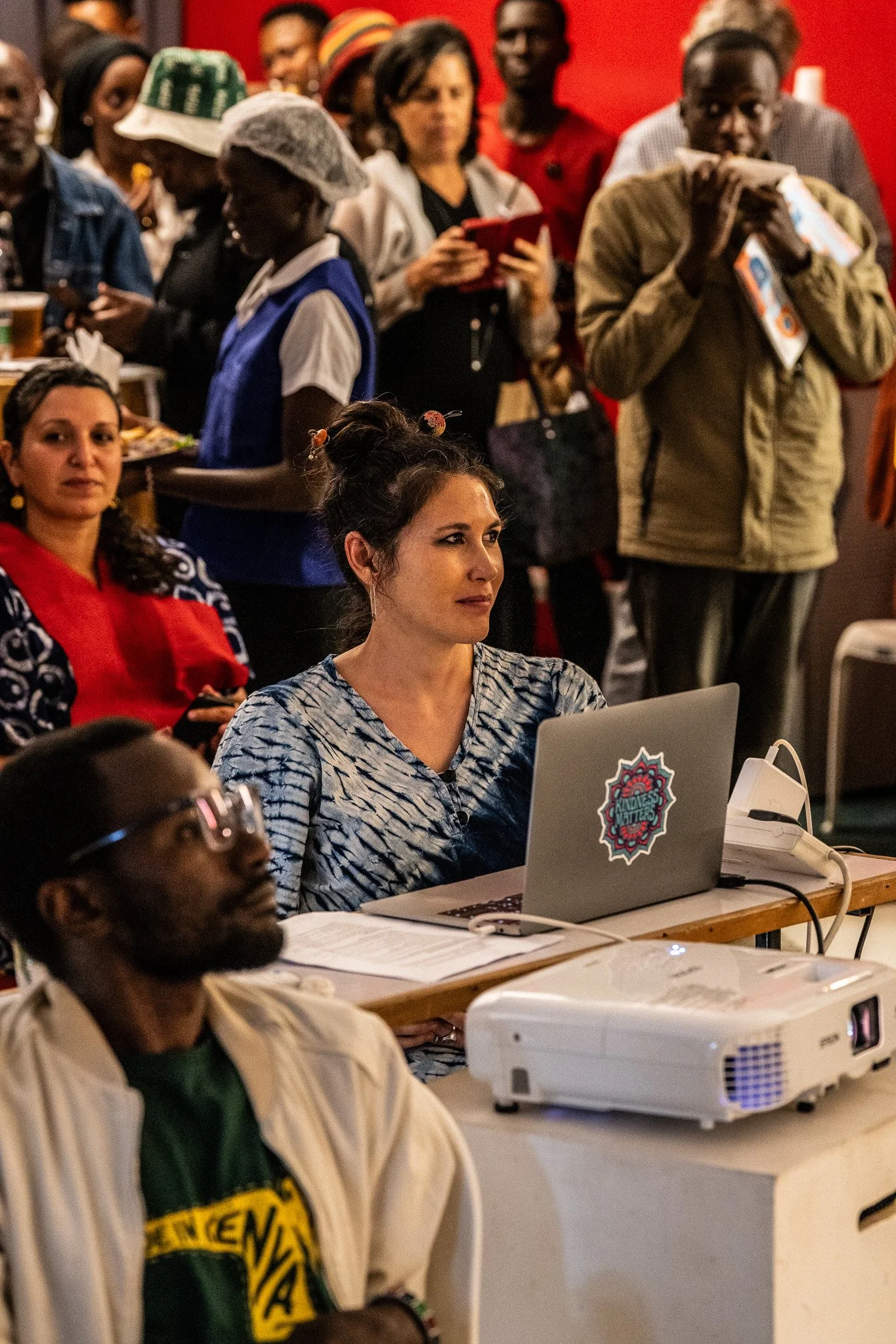 A group of diverse people attending an indoor event, with some seated at a table with a laptop and a projector, while others stand in the background, some using their phones.