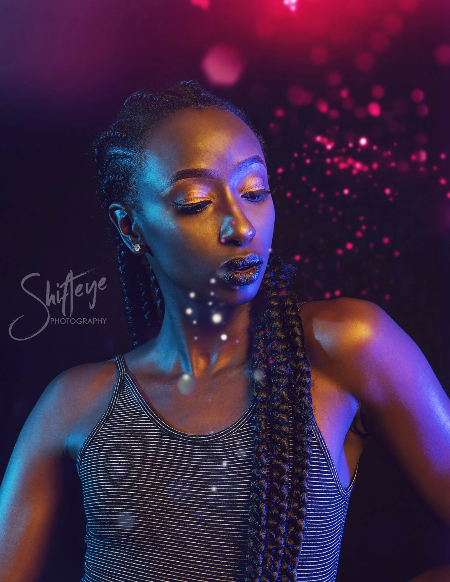 A woman with braided hair and gold makeup poses against a colorful, bokeh-filled background with pink, purple, and blue hues.