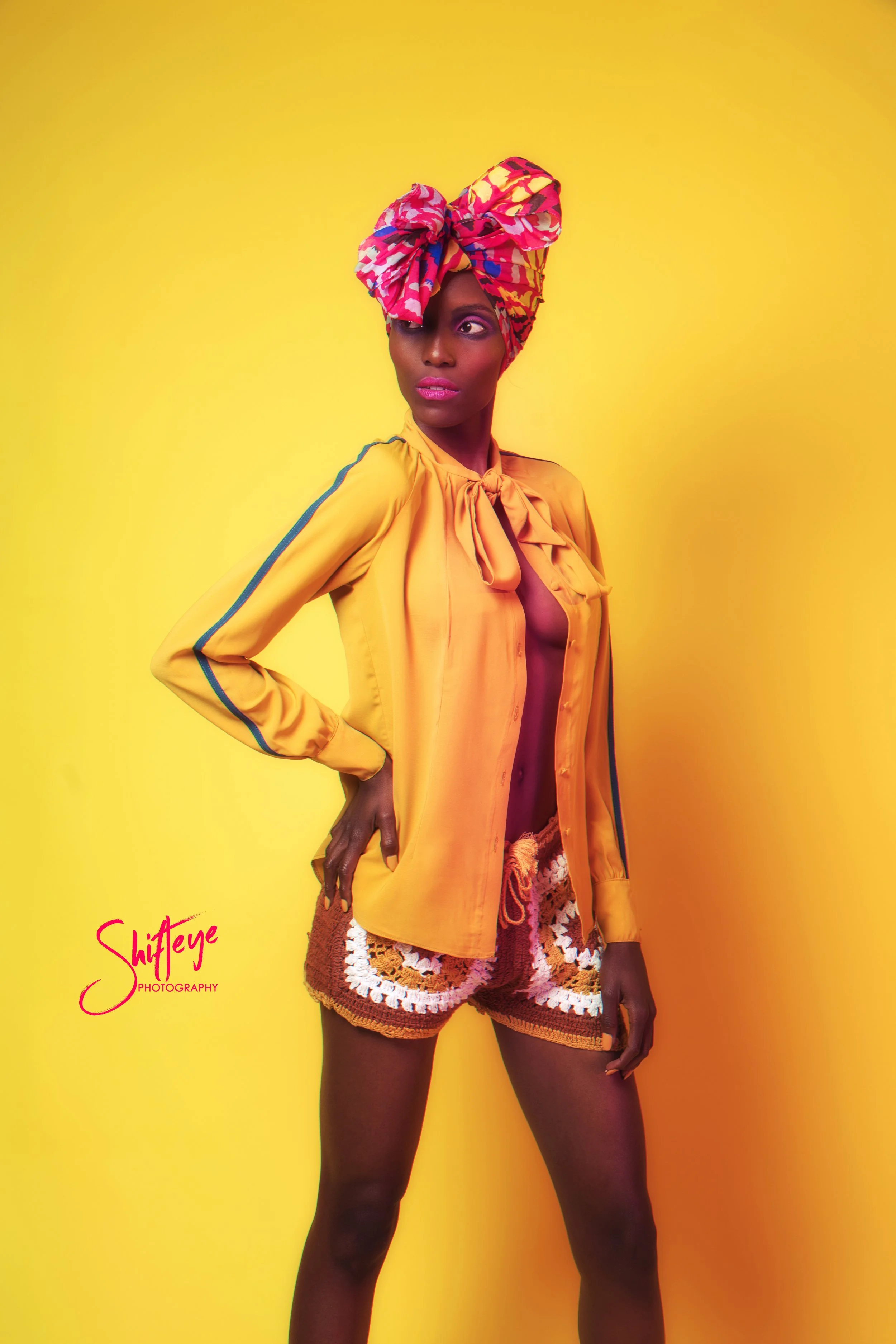 A woman with dark skin in bright yellow background, wearing a colorful headwrap, orange blouse, and crochet shorts.