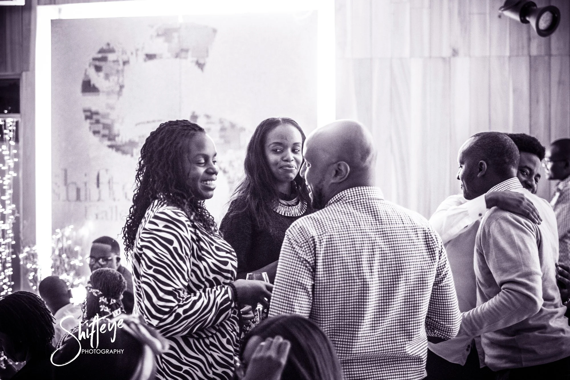 Group of people at a social gathering, smiling and engaging with each other, in a decorated venue with a large portrait in the background, in black and white.