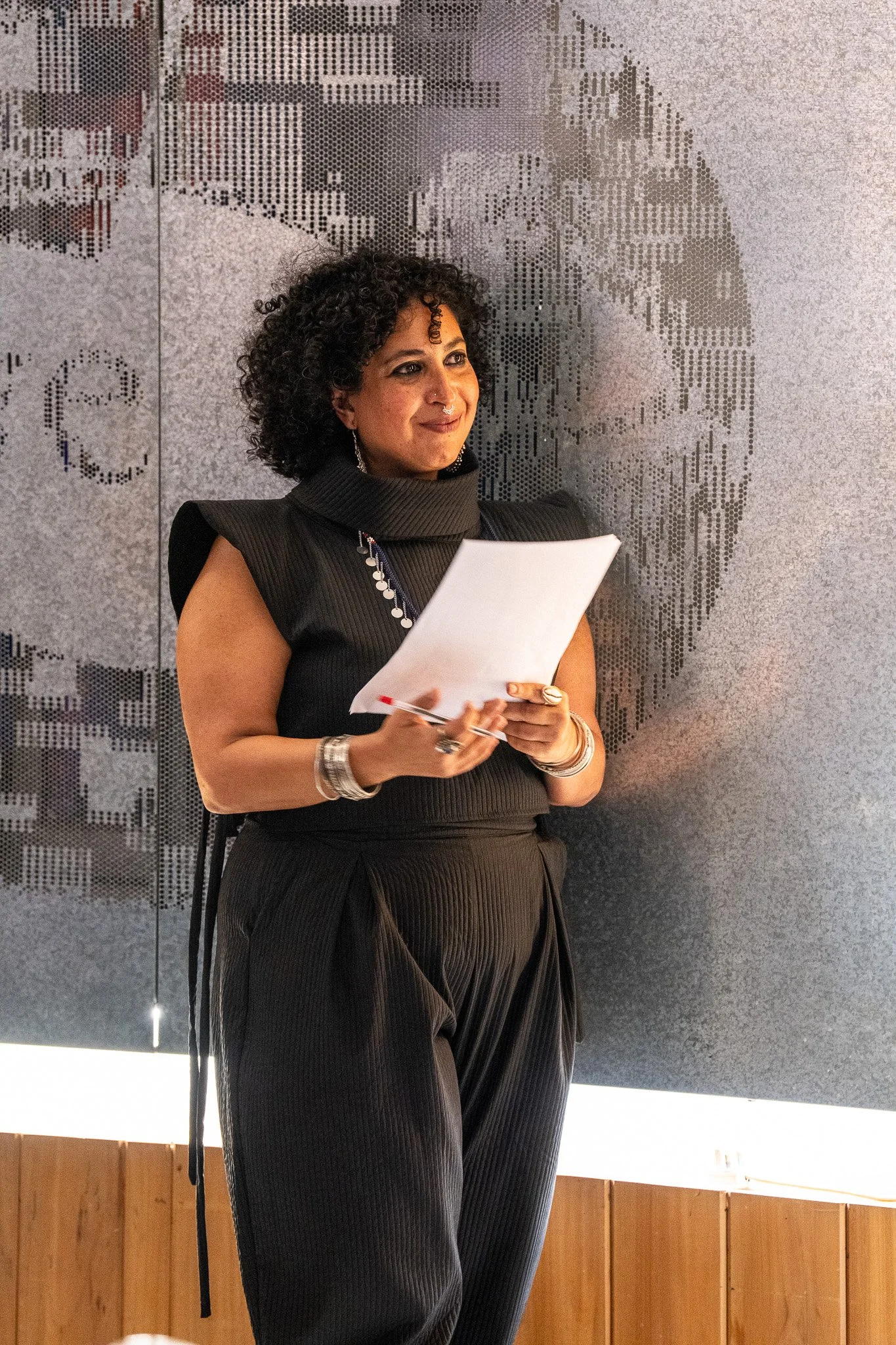 A woman with curly hair standing in front of a wall with a pixelated portrait of a man. She is holding a piece of paper and a pen, smiling slightly, dressed in black with jewelry.