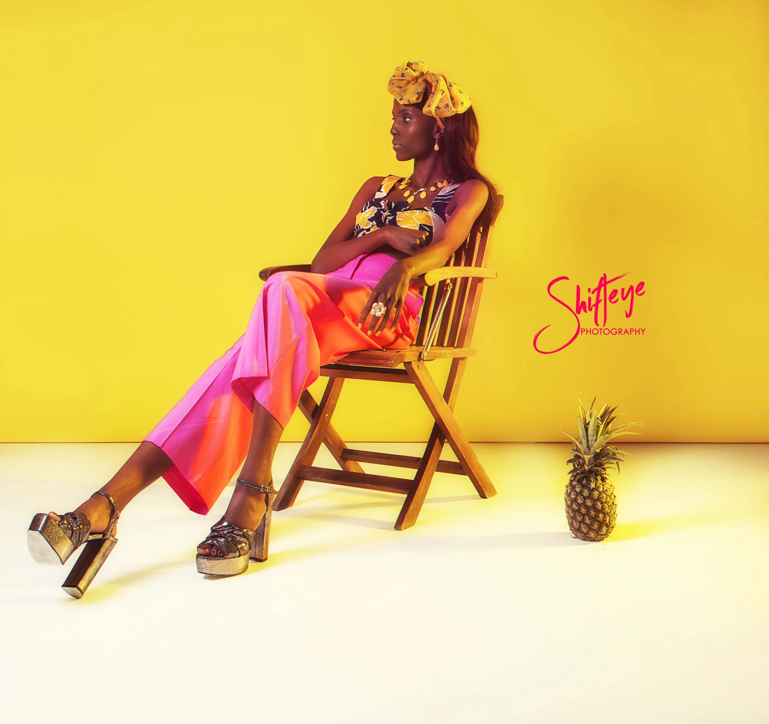 A woman in colorful clothing sitting on a wooden chair with a pineapple on the floor beside her, set against a yellow background. The image includes the text 'Shifeye Photography' in pink.
