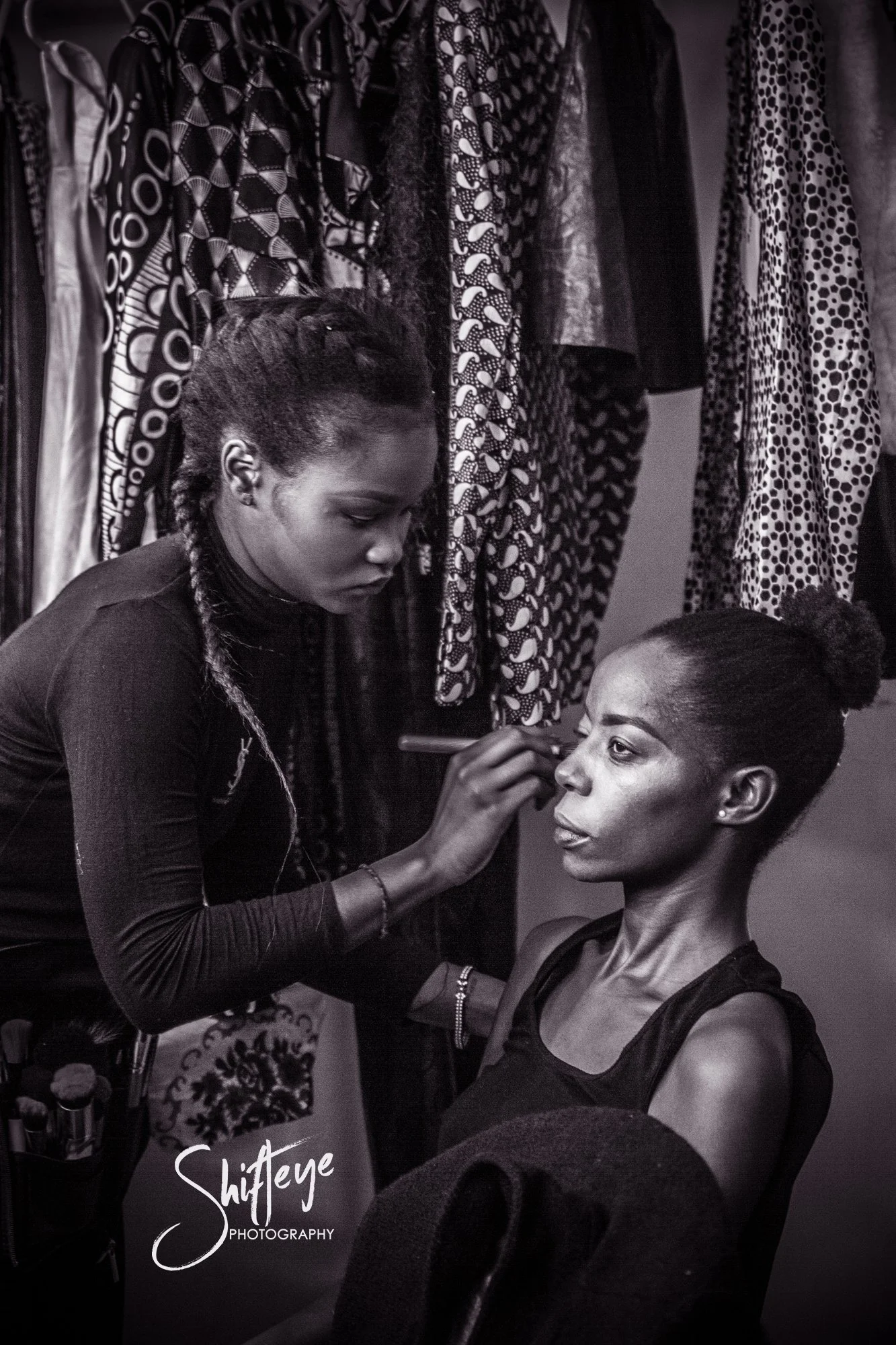 A woman is applying makeup on another woman who is seated, in front of a clothing rack with various patterned garments.