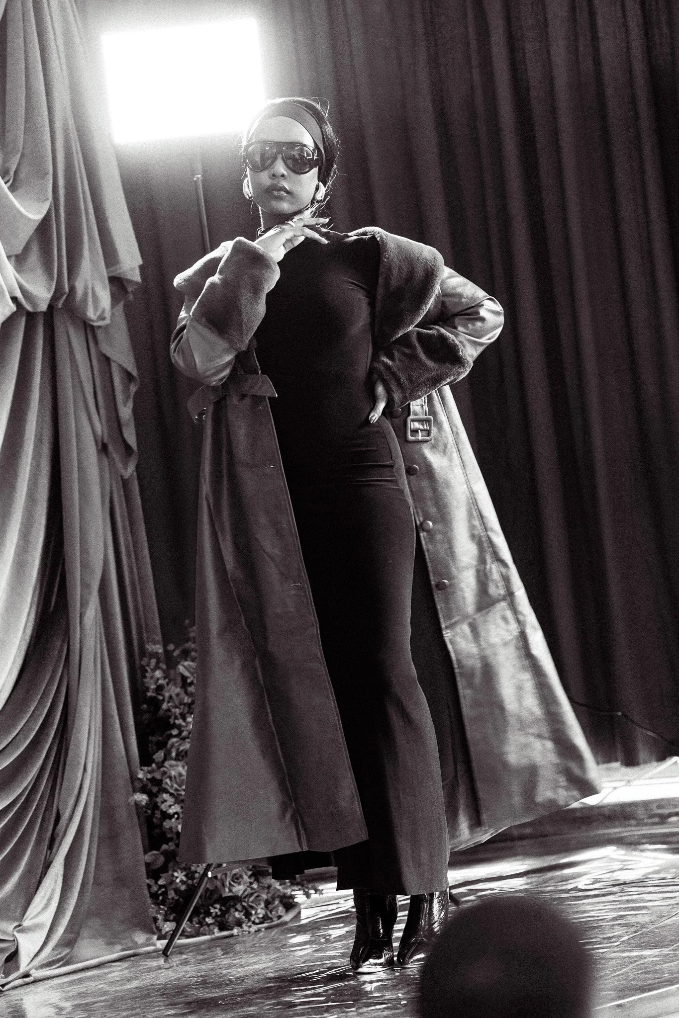 A woman in large sunglasses and a headband posing confidently on a stage, wearing a long coat and high heels, with curtains and stage lighting in the background.