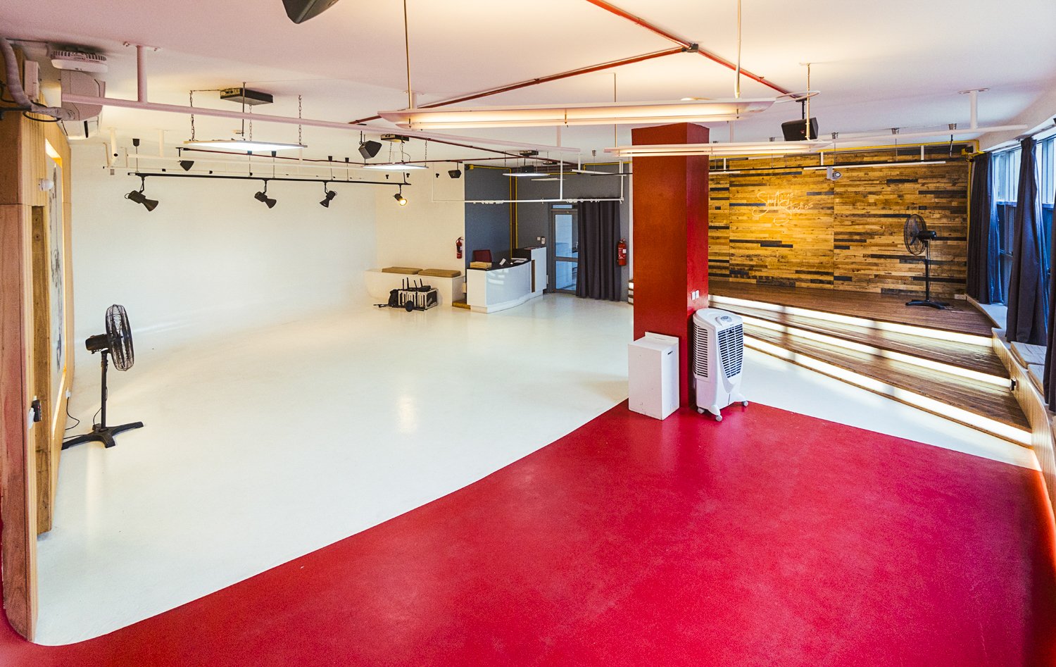 Empty dance studio with red and white flooring, wooden wall, stage, black curtains, stage lighting, and air conditioning units.