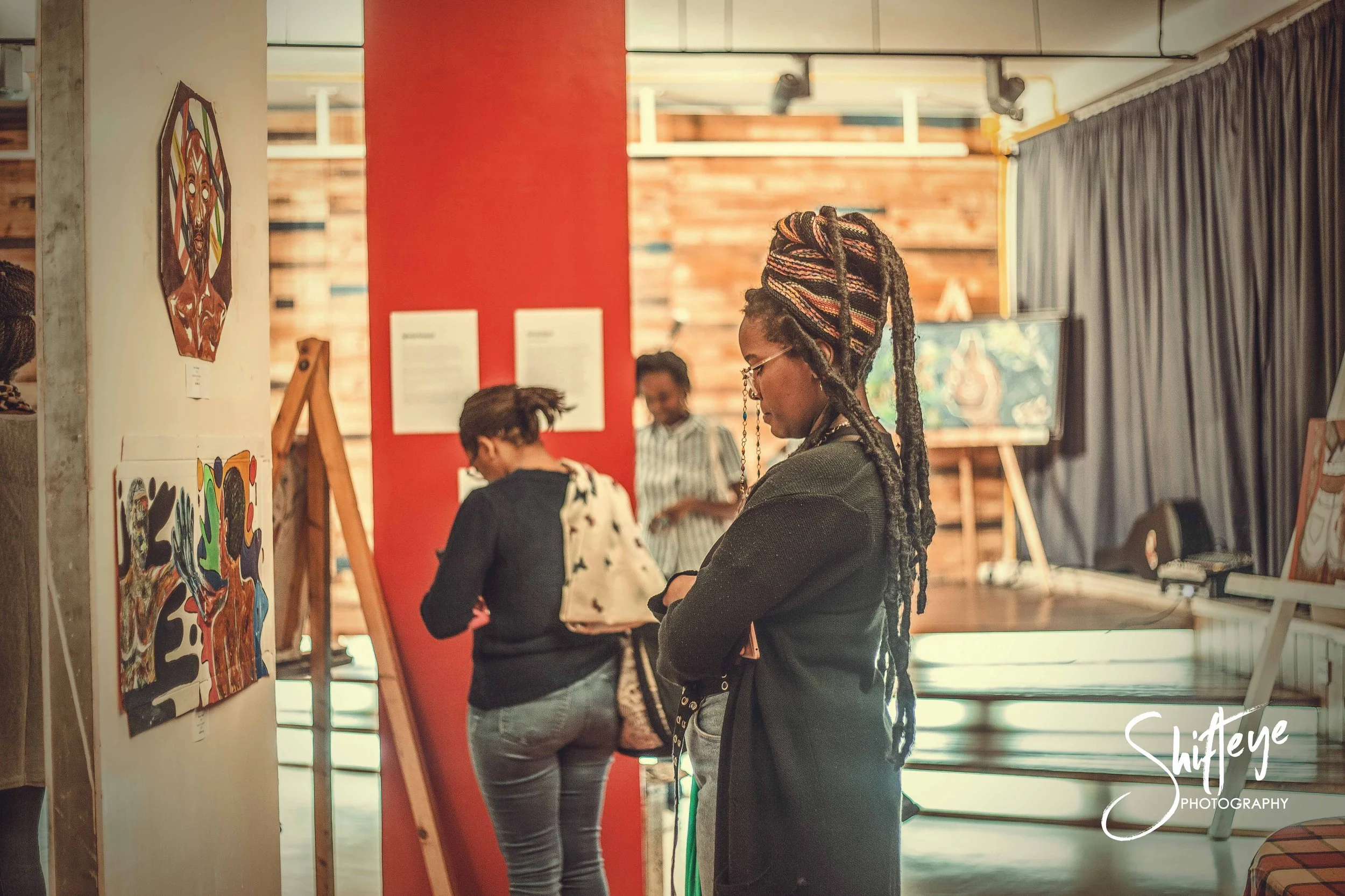 People viewing artwork in an art gallery with paintings on the walls and easels. One woman with dreadlocks and glasses is in the foreground, looking at her phone. Others are in the background, observing art.