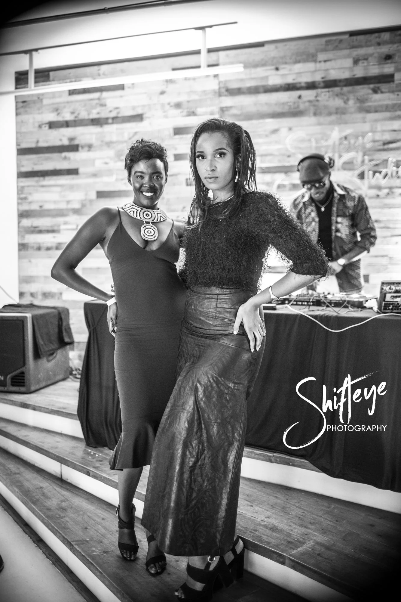 Two women posing together at an event, with a man at a DJ booth in the background. The women are dressed in elegant evening attire, and the setting has a wood-paneled wall. The photo is in black and white.