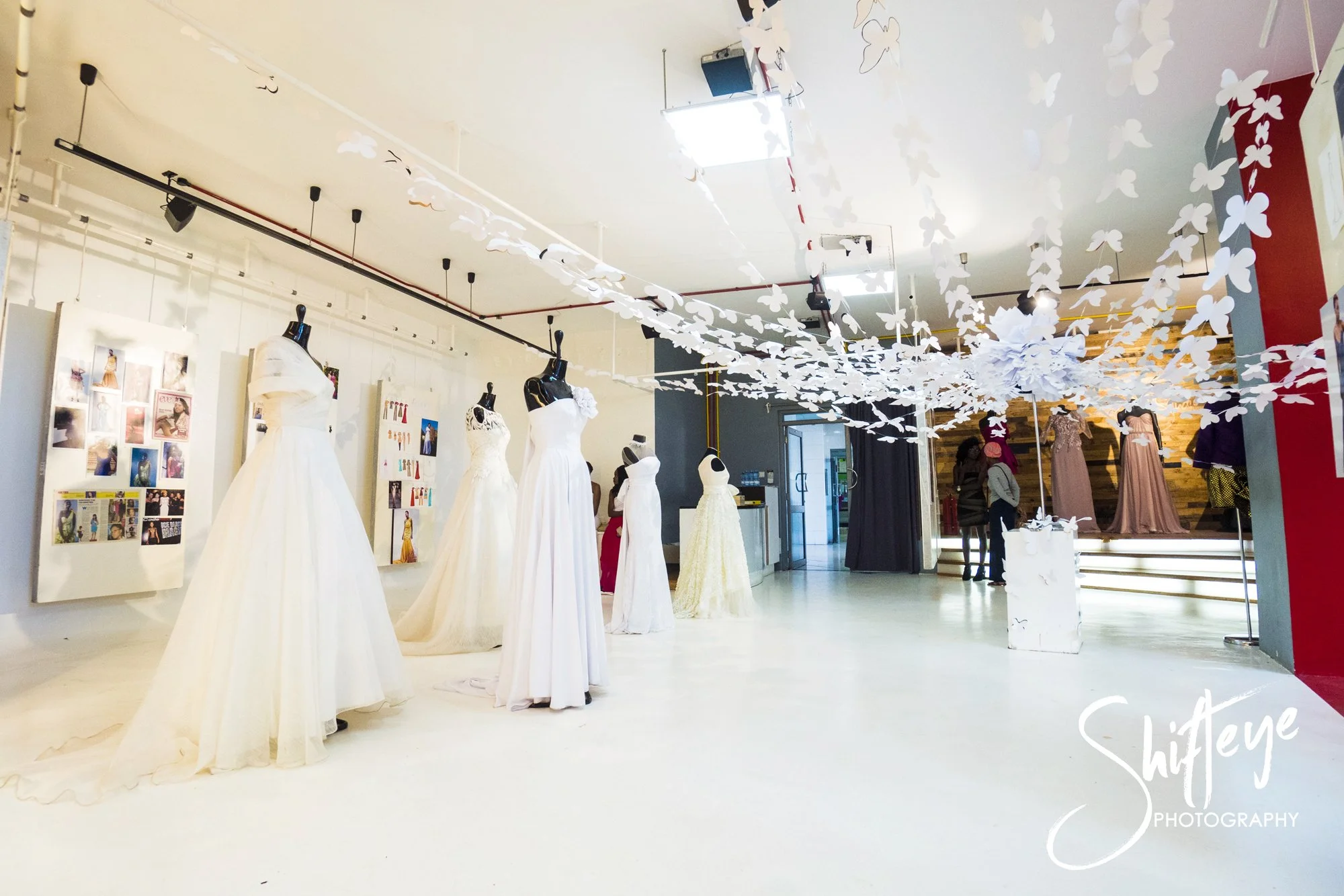 An interior view of a bridal boutique featuring several mannequins dressed in white wedding gowns, decorative white butterfly and flower wall hanging, staircase with gowns on display, and customers browsing.