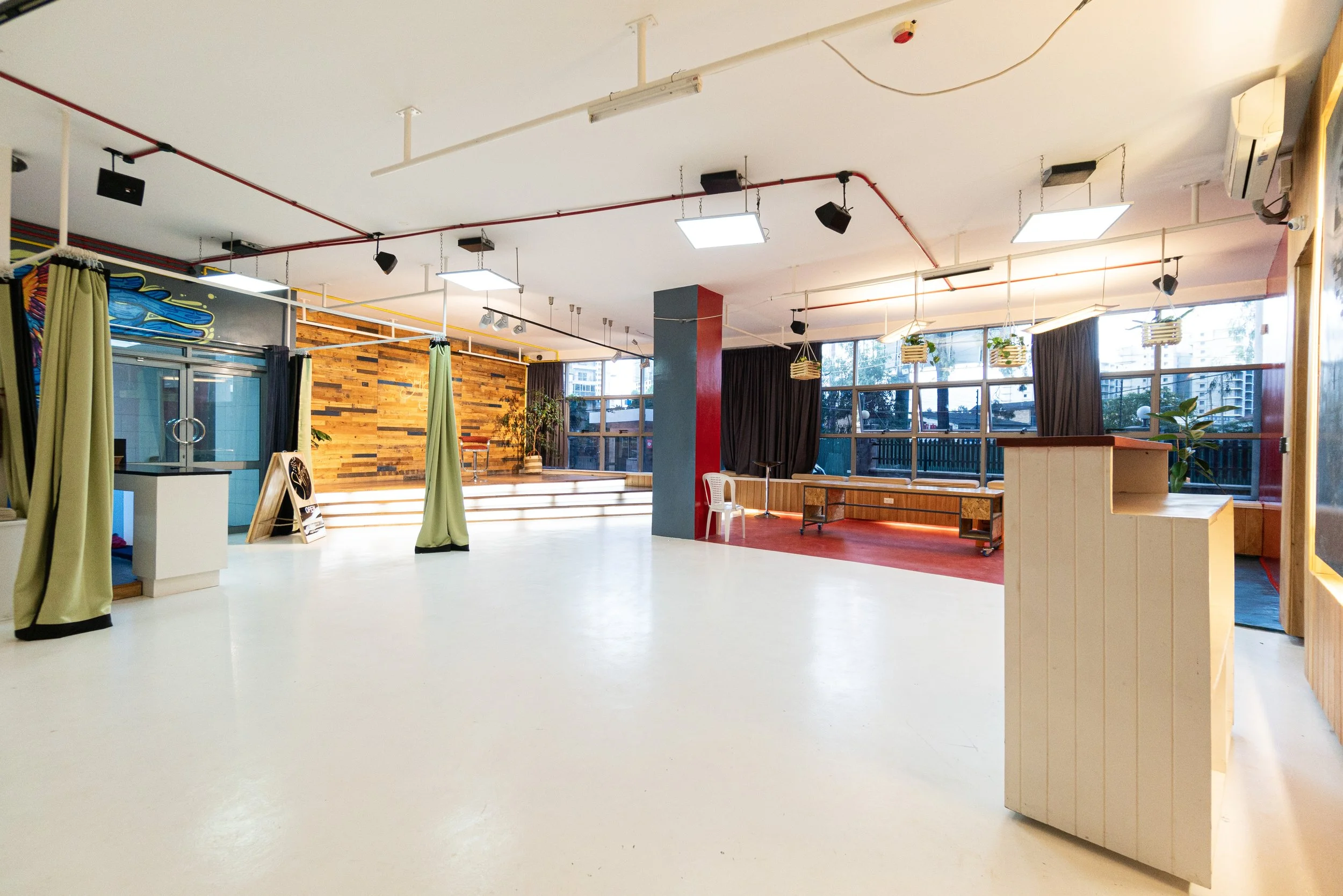 A spacious room with large windows, wooden accent walls, and tiered seating. The floor is mostly white, with a red area rug. There are black curtains, hanging plants, and various lighting fixtures on the ceiling.
