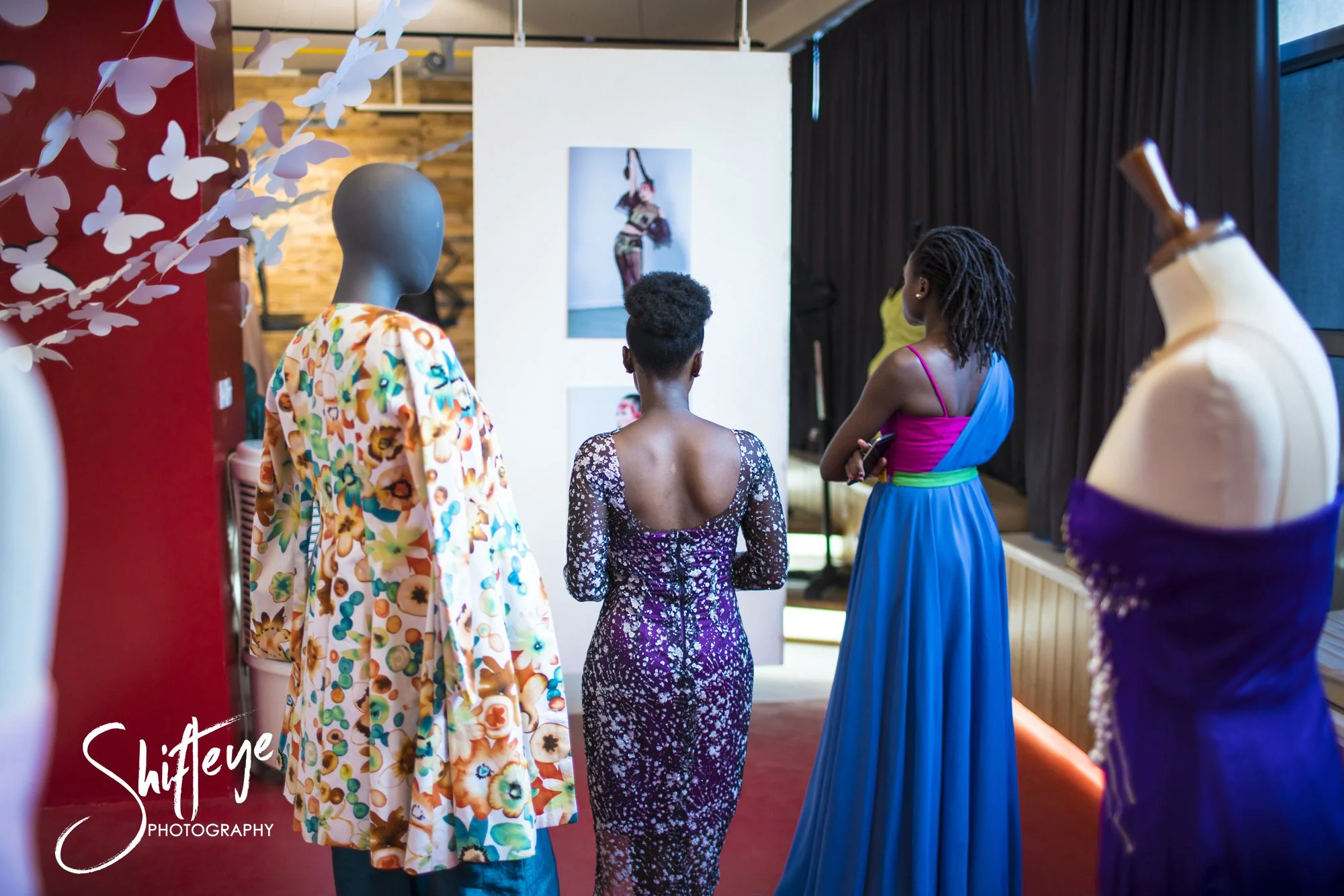 Three women and two mannequins in a dress shop, with dresses on display and a photographer's logo in the bottom left corner.