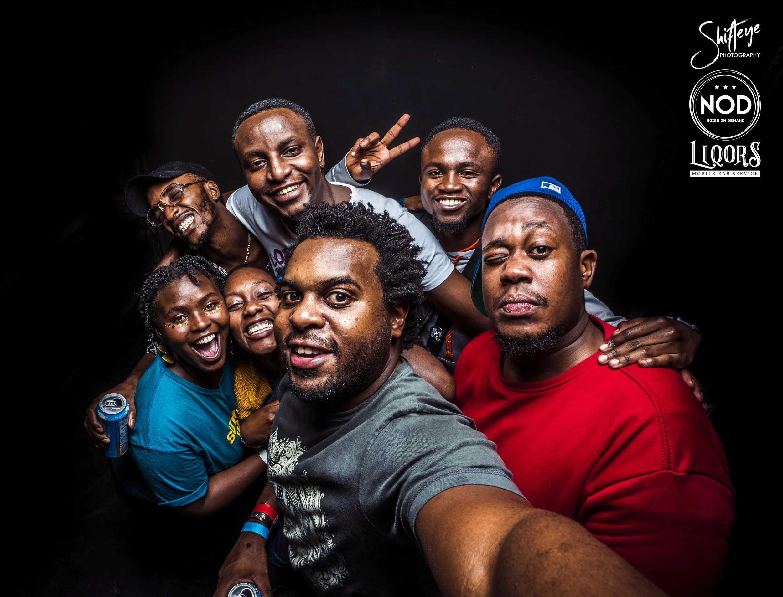 A group of seven cheerful people taking a selfie together against a black background, with some smiling, laughing, and making peace signs. Logos in the top right corner indicate photography and bar service branding.