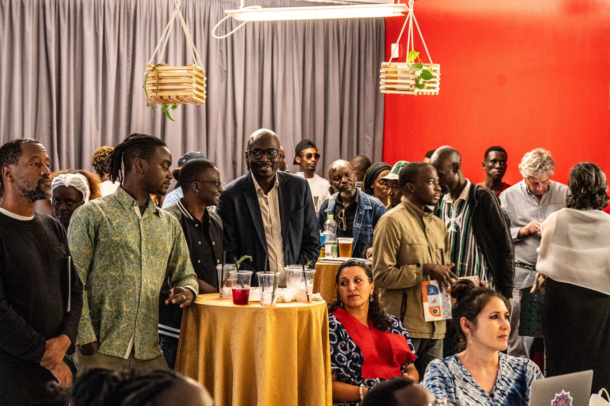 A diverse group of people gathered at an indoor event, some standing around a yellow table with drinks and others seated. The room has gray curtains and red walls, with hanging wooden planters and a fluorescent light fixture.