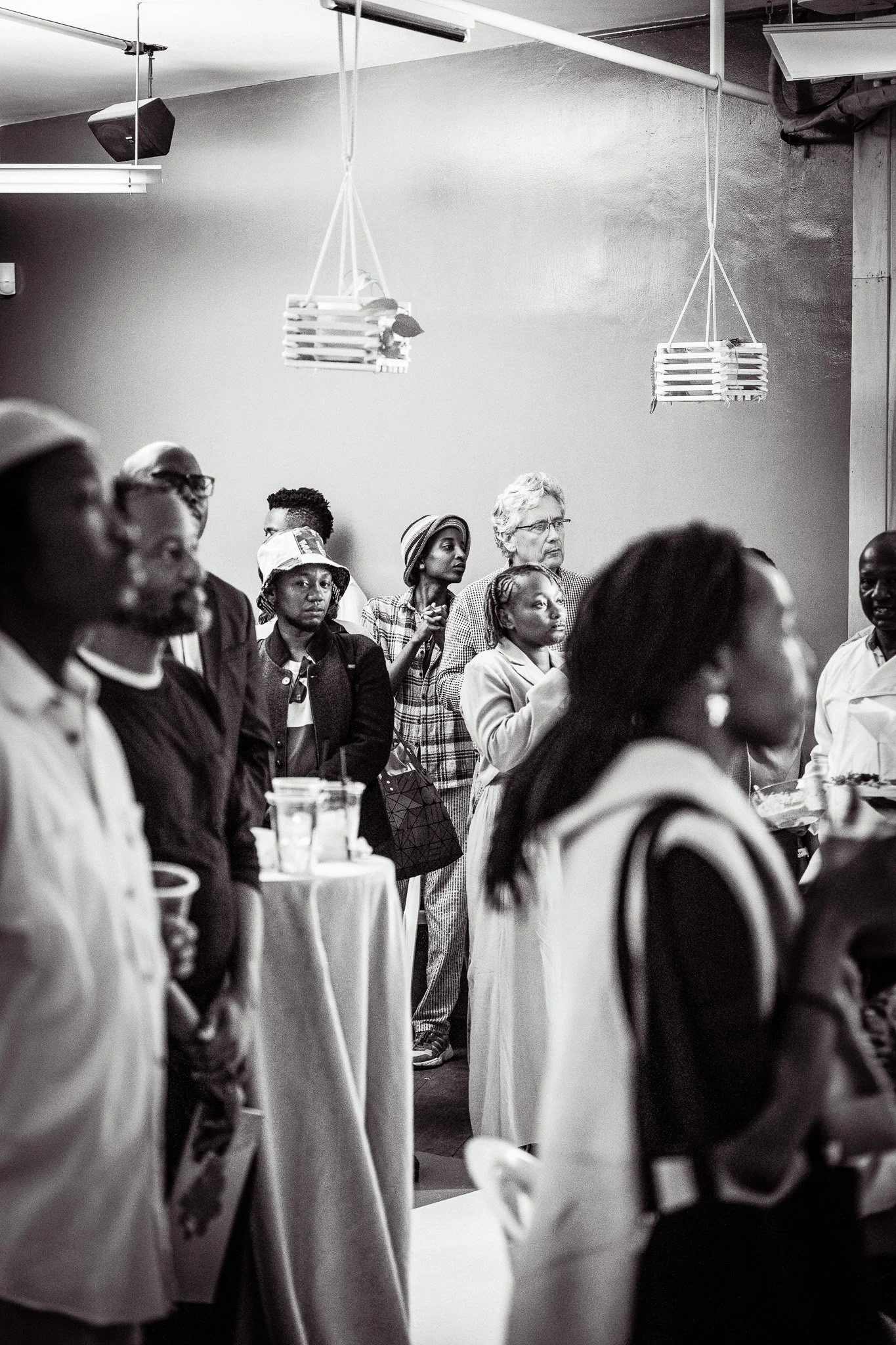 A black and white photo of a diverse group of people standing and listening attentively in what appears to be an indoor event space or gallery.
