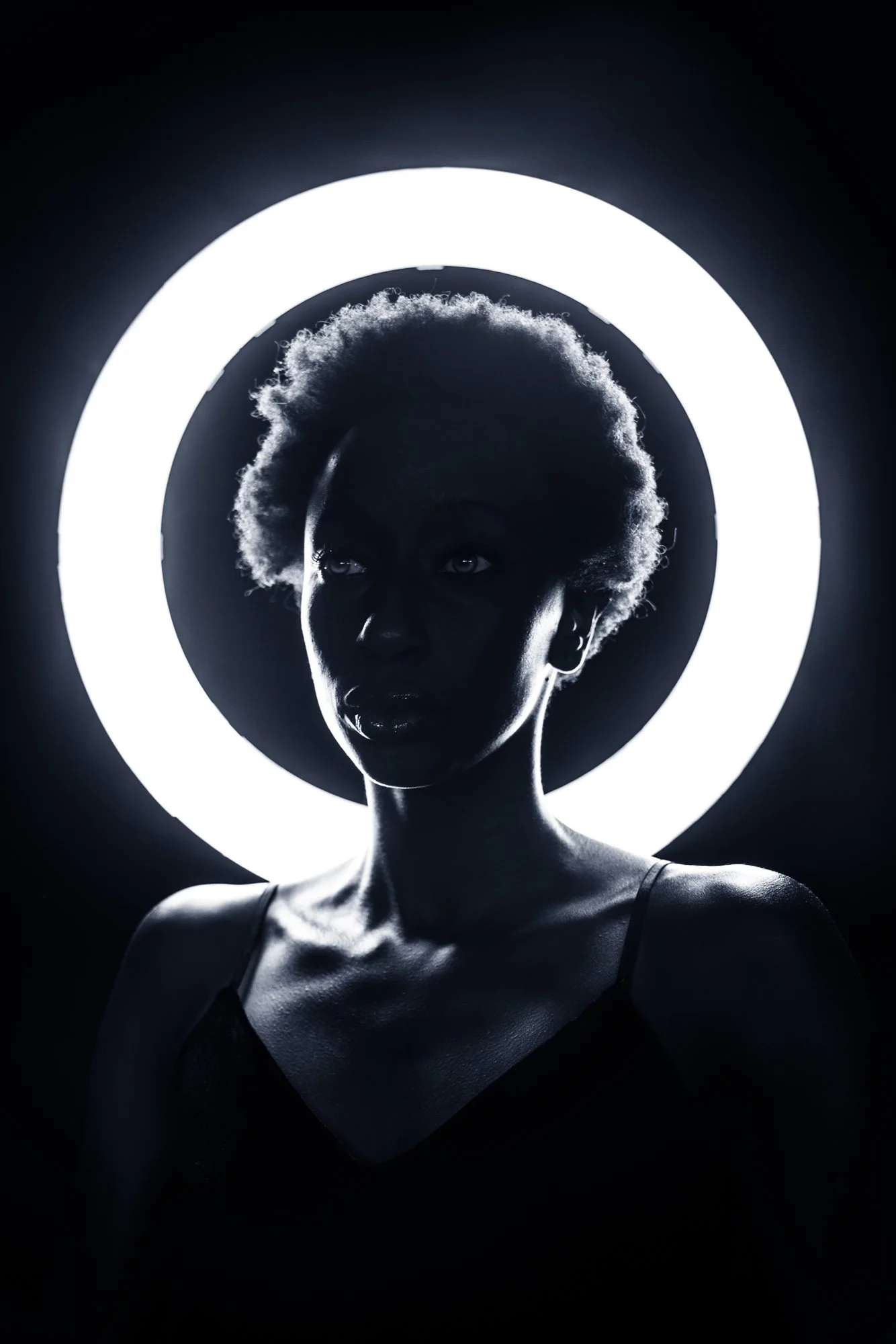 A black and white photo of a woman with natural hair, illuminated by a bright circular light ring behind her, creating a silhouette and dramatic lighting effect.
