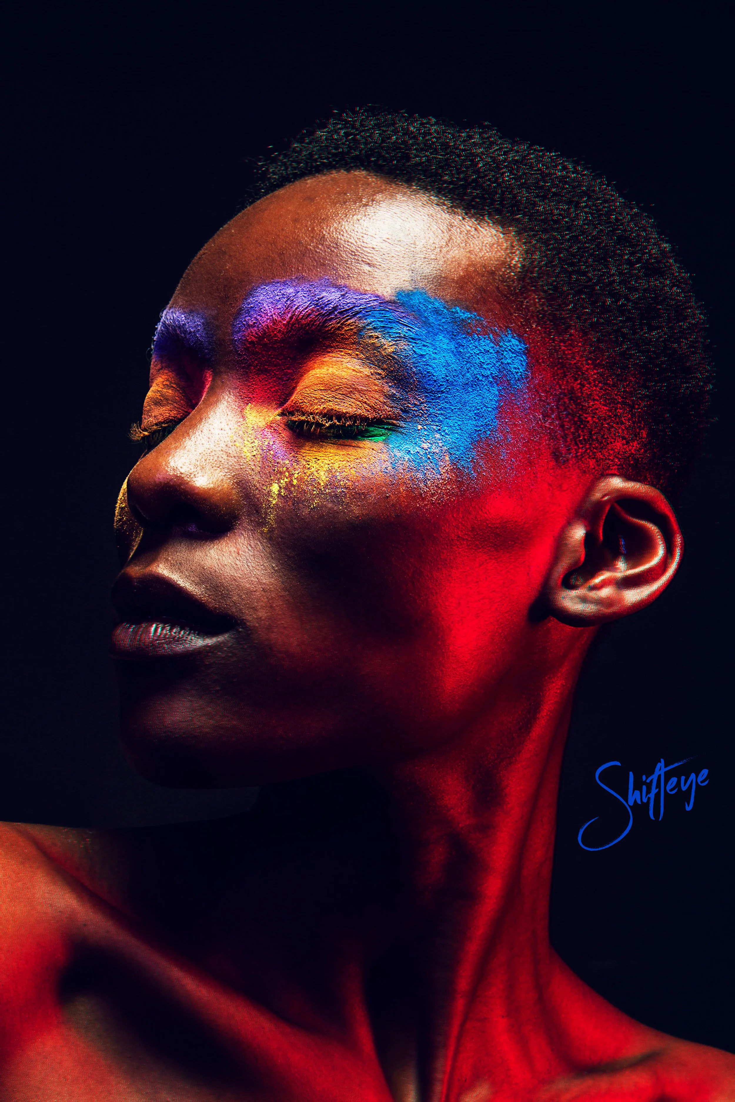 Close-up of a woman with her eyes closed, face painted with vibrant colors including blue, red, yellow, and purple, against a black background.