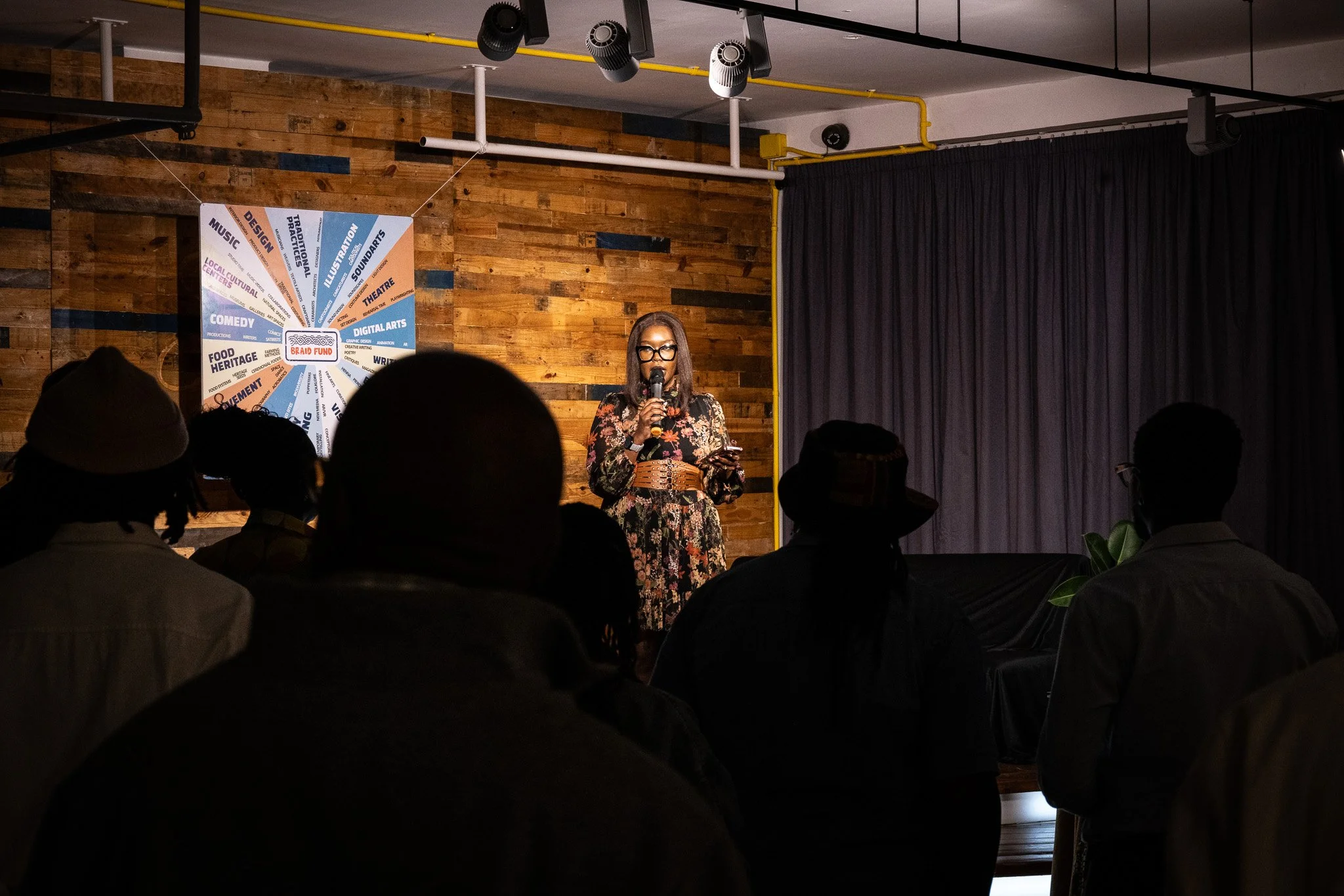 A woman speaking into a microphone on a stage with a dark curtain background, in front of an audience at an indoor event space with wooden walls and ceiling lights.
