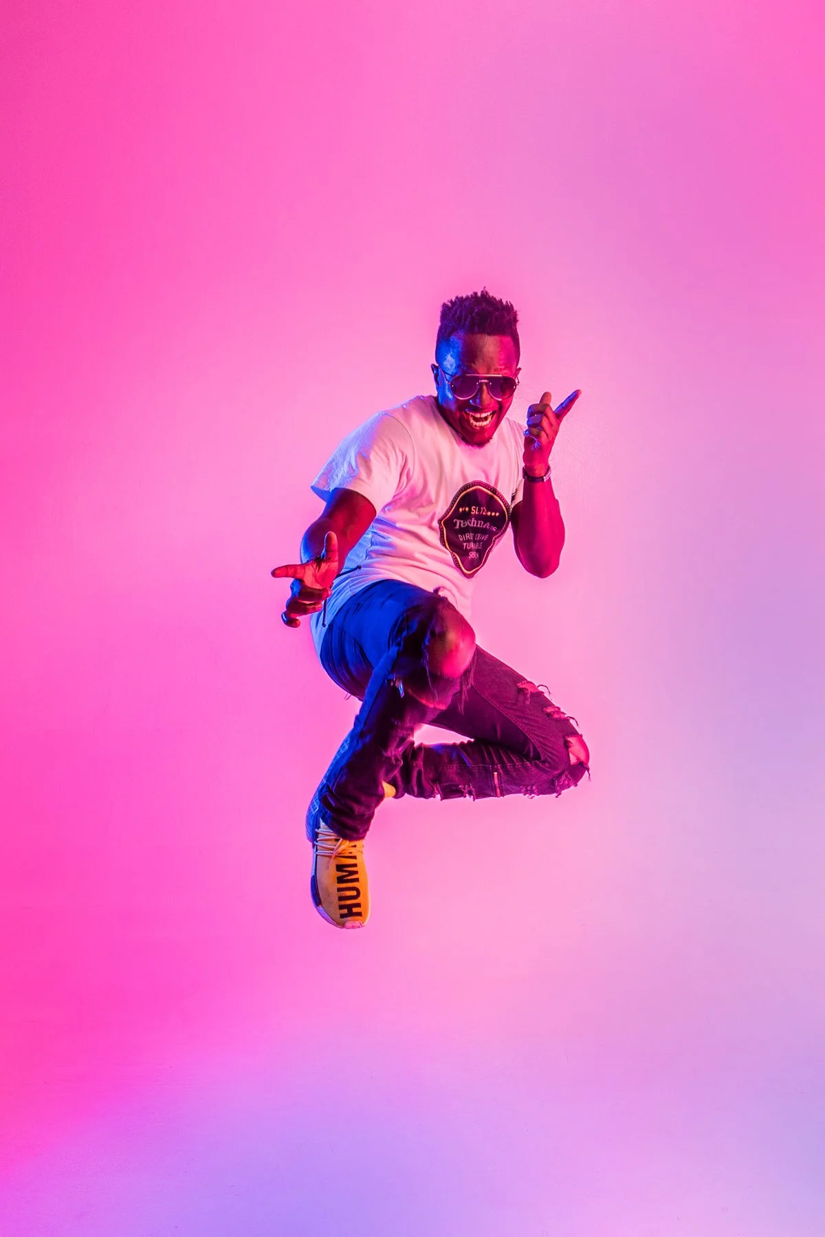 A man with sunglasses jumping against a pink and purple gradient background, wearing a white t-shirt, ripped jeans, and yellow shoes with 'HUMAN' written on the sole.