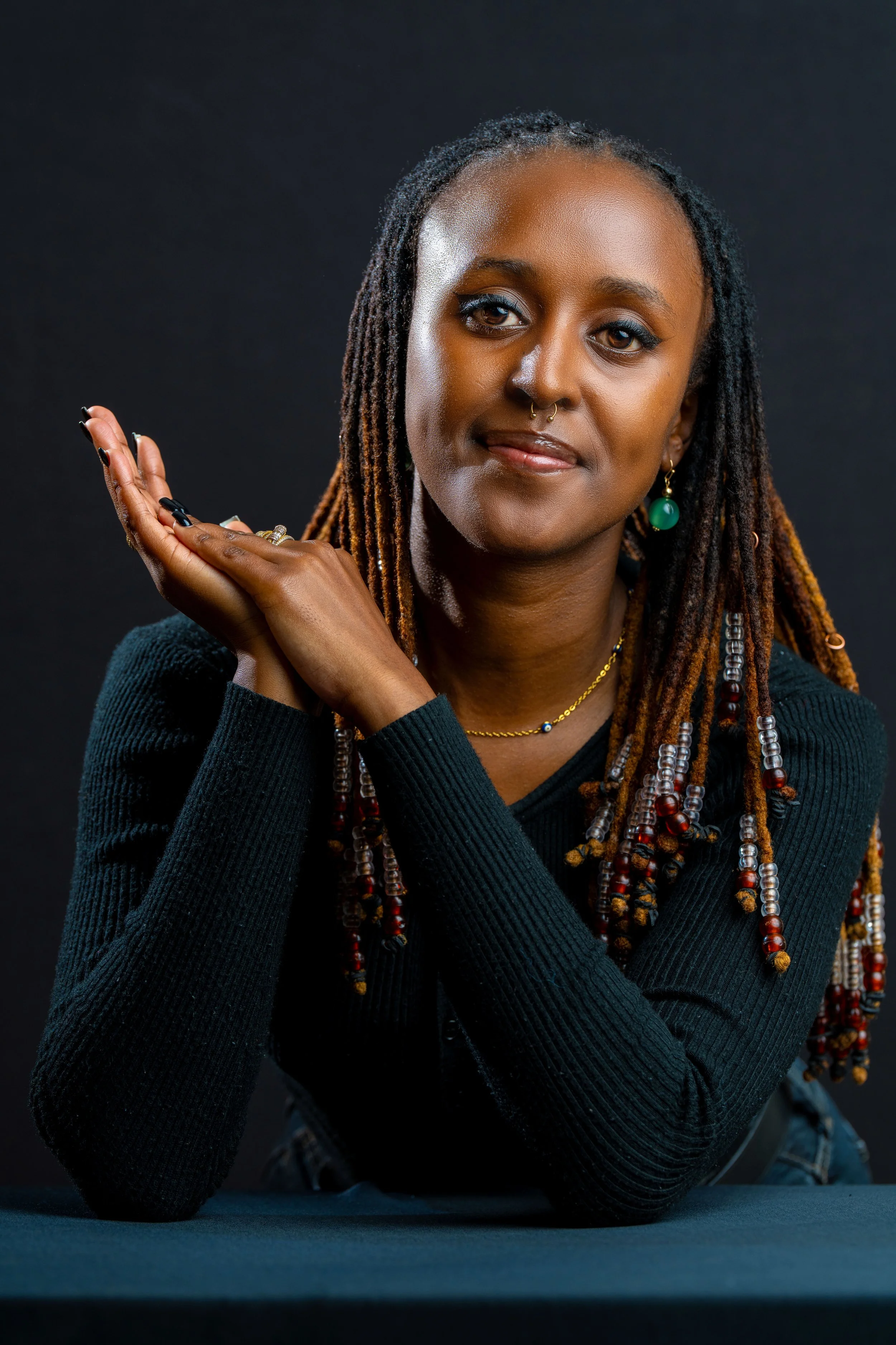 Portrait of a woman with dreadlocks, wearing a black long sleeve top, gold jewelry, and green earrings, posed with hands near her face against a dark background.