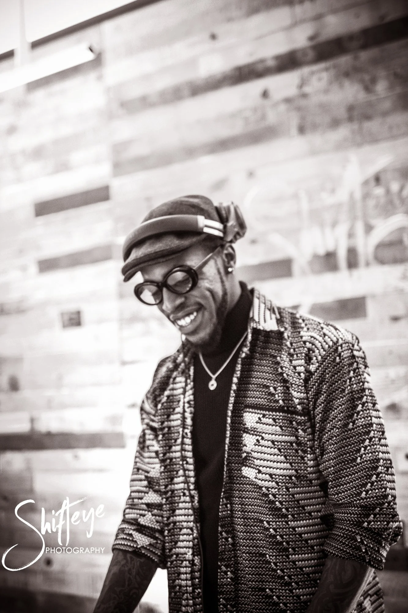 A smiling man wearing glasses, headphones, a hat, and a patterned jacket standing in front of a wooden wall.