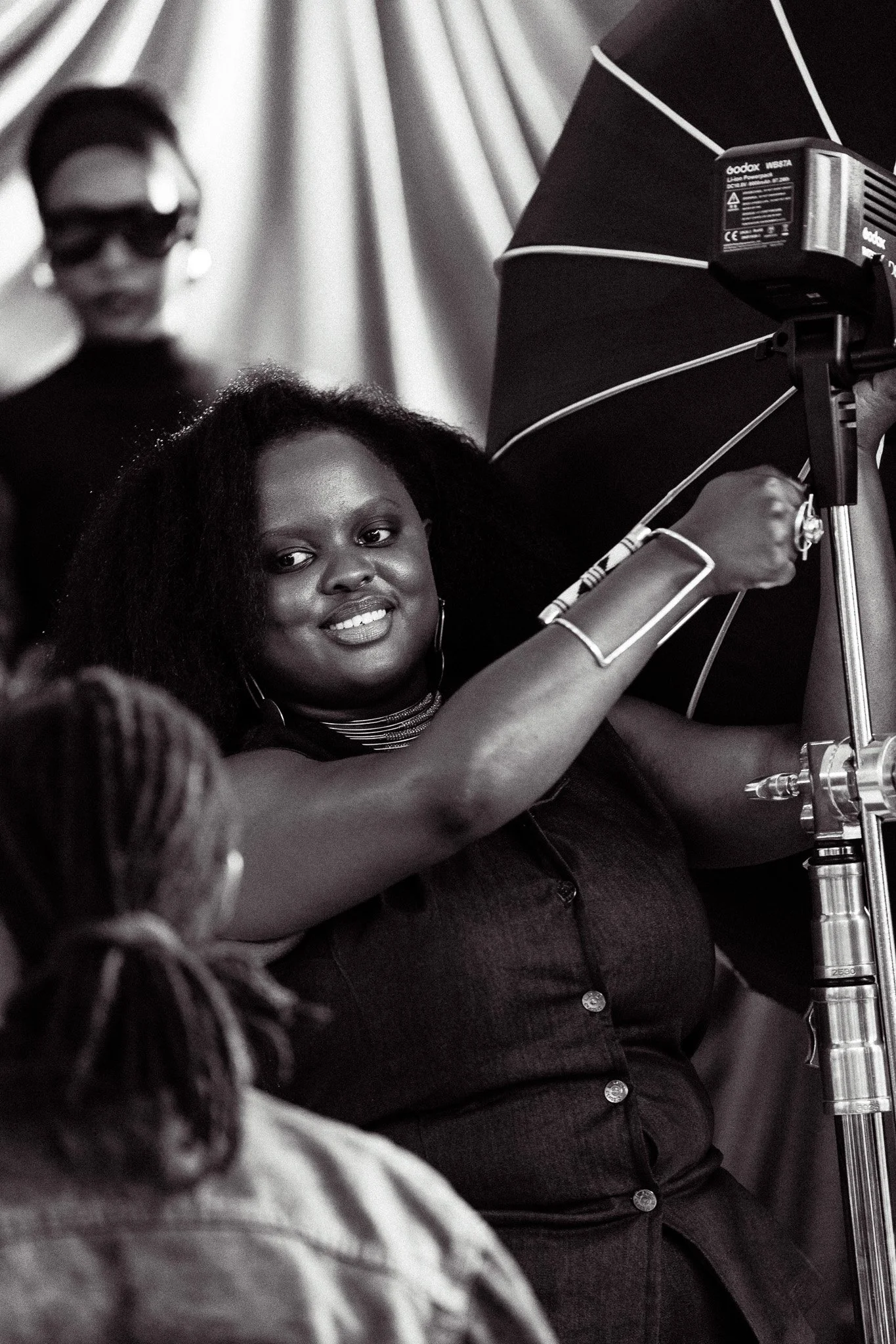Black woman adjusting a studio light on a photography set, smiling at the camera, with another woman and a person with long hair in the background.